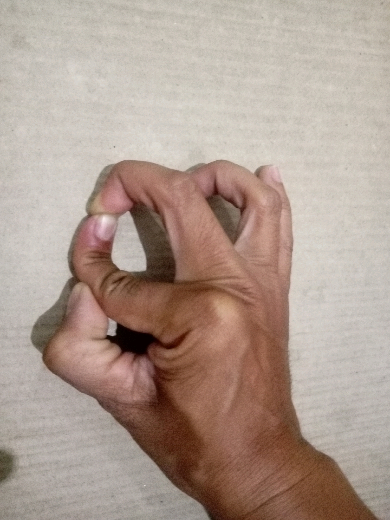 A hand is positioned in front of a light-colored, textured surface, possibly a wall or floor. The hand is making a gesture where the thumb and index finger are forming a circle, and the other fingers are bent inwards. The skin tone of the hand is medium to dark brown. The lighting casts a subtle shadow to the left of the hand. The image does not contain any elements that would indicate a specific location within Batam, Indonesia.