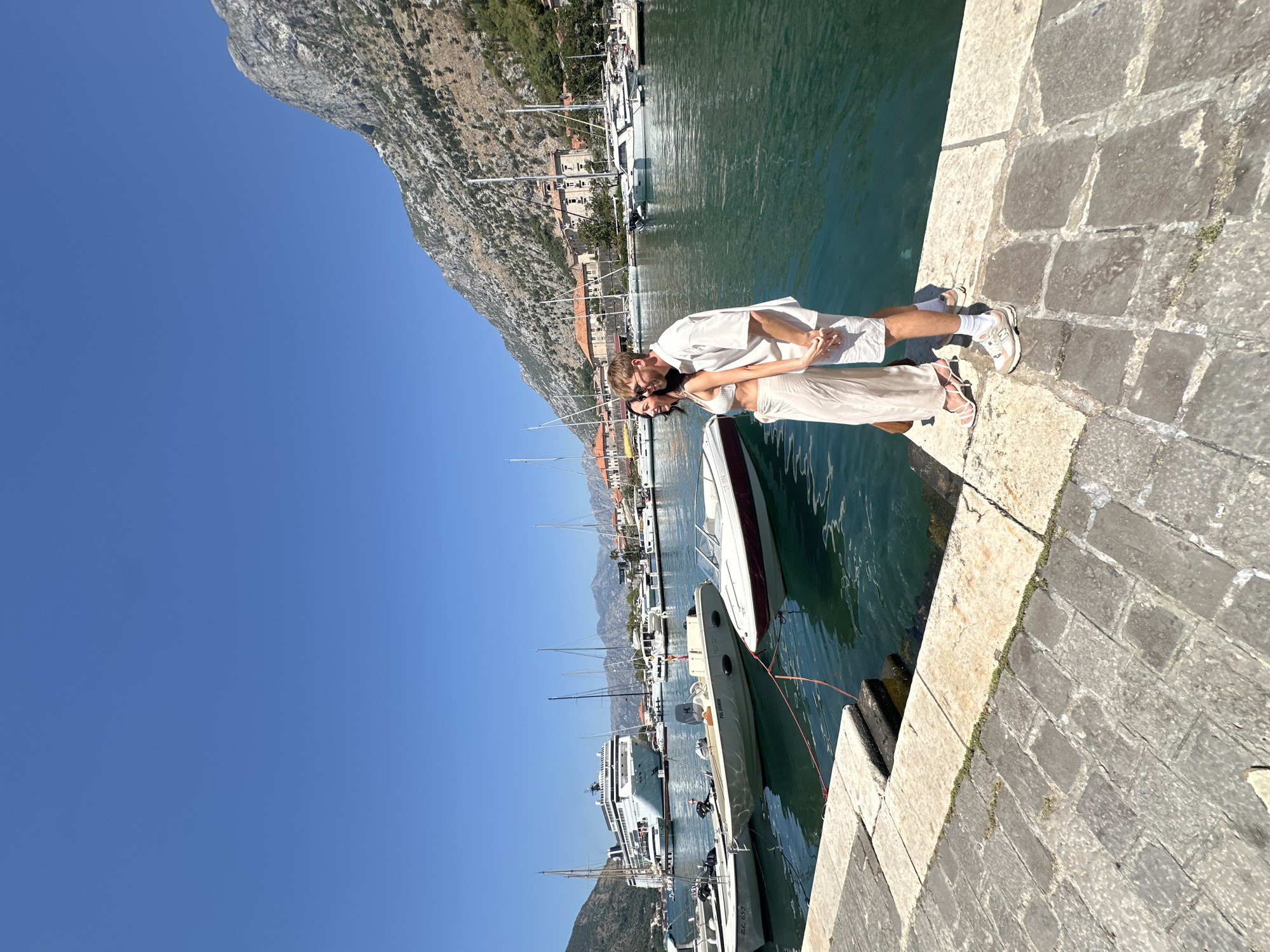 In the city of Kotor, Montenegro, a couple stands on a stone quay, holding hands and looking out at a harbor filled with boats. The man, wearing a light-colored short-sleeved shirt and white shorts, has his arm around the woman, who is dressed in a cream-colored crop top and flowing pants. In the background, a large cruise ship is docked, along with numerous sailboats and smaller motorboats. The water is a vibrant turquoise, reflecting the clear blue sky and the rugged, tree-covered mountains that rise steeply behind the city's buildings. The stone quay is paved with large, irregular flagstones, and a few tufts of grass sprout from the cracks between them. The scene is bathed in bright sunlight, indicating a warm, sunny day.