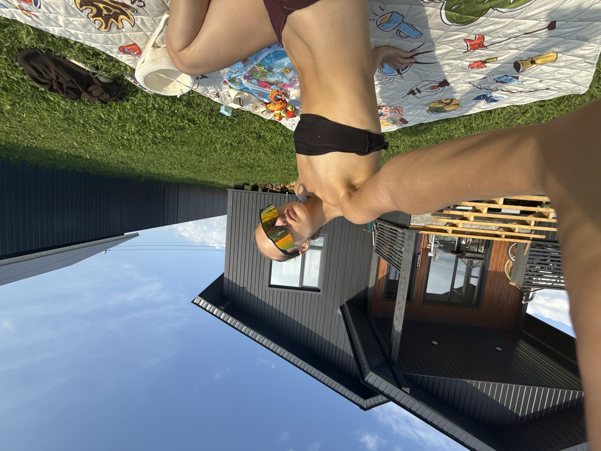 A young woman is taking a selfie in a backyard in the village of Podshivalovo, Russia. She is wearing a black bikini top and maroon bottoms, with large, reflective sunglasses. She is sitting on a patterned blanket on the grass. In the background, a modern, dark-colored house with wooden accents is visible. The sky is blue with some scattered clouds. There are also some children's toys on the blanket, and a white woven basket. A dark garment is lying on the grass to the right of the blanket. The overall scene suggests a leisurely day outdoors.