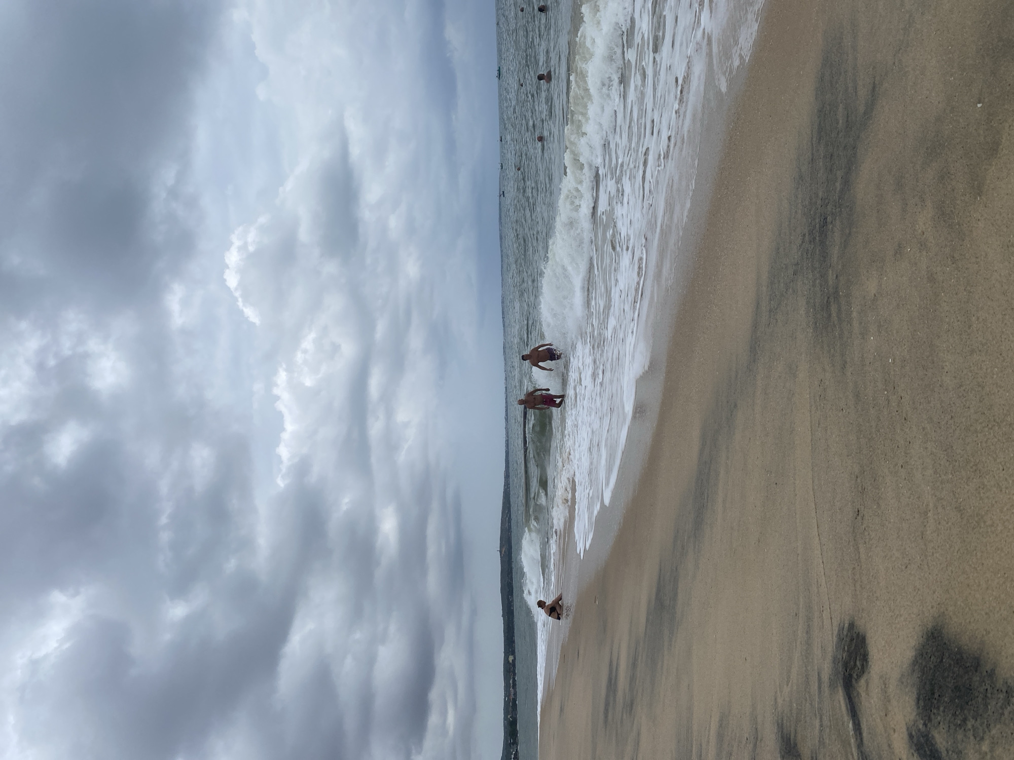 This image captures a beach scene in Phu Thuy Ward, Vietnam, on an overcast day. The composition features a broad expanse of sandy shore on the right, the ocean with waves on the left, and a cloudy sky dominating the upper portion.
Several people are visible in and around the water. Two adult men are standing in the shallow ocean water, with waves breaking around them. One man, wearing light-colored shorts, is closer to the viewer, partially obscured by a small wave. Another man is positioned further to the left, also in the water. Closer to the shoreline, a smaller individual is seated or crouching in the wet sand where the waves recede. Additionally, multiple heads are visible further out in the water, indicating more people swimming or bathing.
The ocean displays moderate waves with white foam along the surf line. The sand is light brown with notable dark, irregular patches, possibly wet sand or mineral deposits. The sky is heavily clouded with various shades of grey, indicating an overcast or potentially stormy day. A faint outline of a distant landmass or coastline is visible on the horizon beneath the clouds. The overall scene suggests a recreational day at the beach despite the cloudy weather. No visible text is present in the image.
