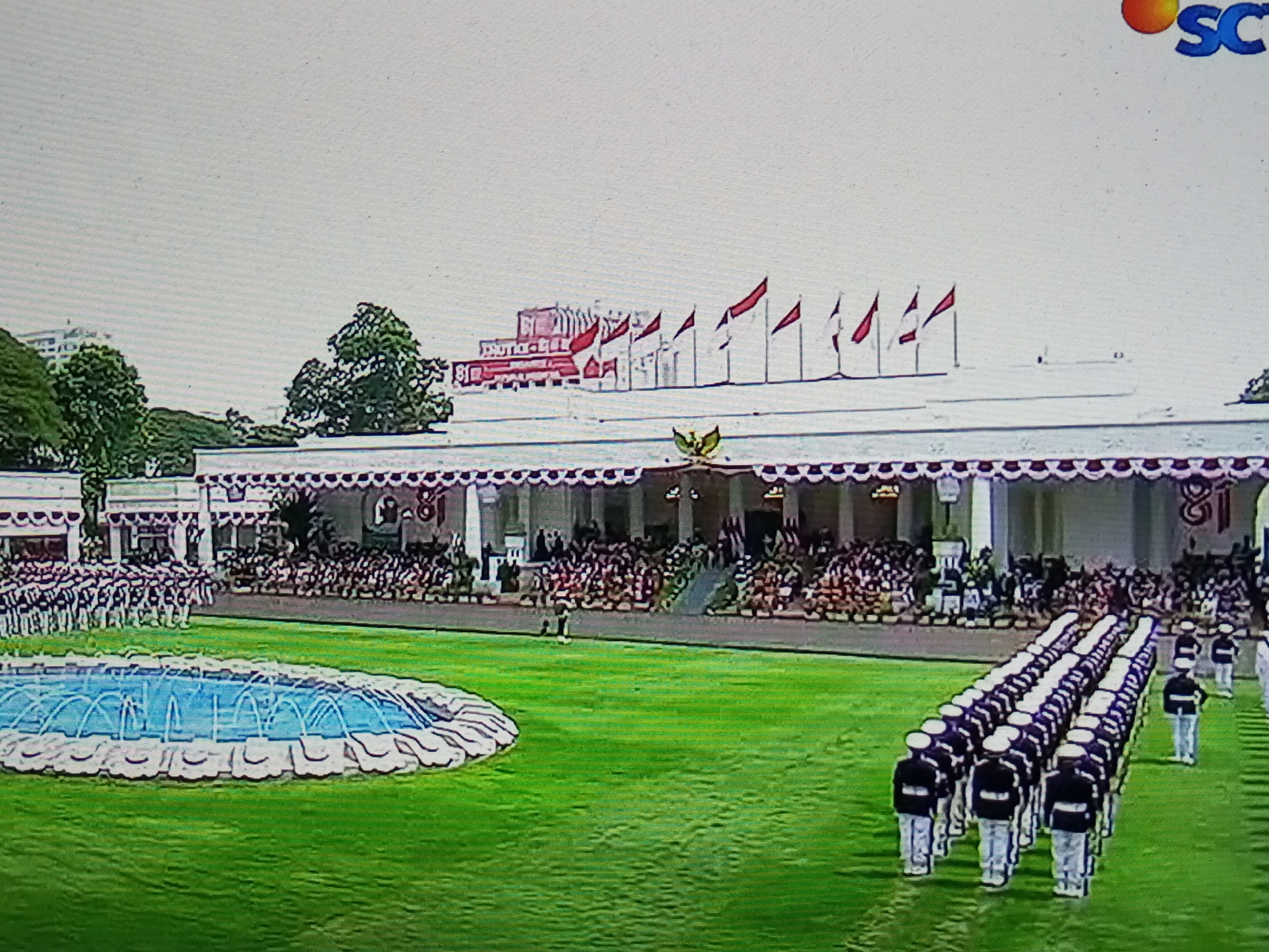 In the city of Banyudono, Indonesia, a formal outdoor ceremony is taking place on a lush green lawn. A large, white, colonial-style building with decorative awnings serves as the backdrop. In front of the building, a crowd of spectators is seated, their faces obscured by the distance. A uniformed marching band, comprised of individuals in dark jackets and white pants, is positioned on the lawn, their formations neat and orderly. In the foreground, a decorative fountain with blue water sprays is visible. Several red and white flags are flying from poles in front of the building. The overall scene suggests a national event or celebration.