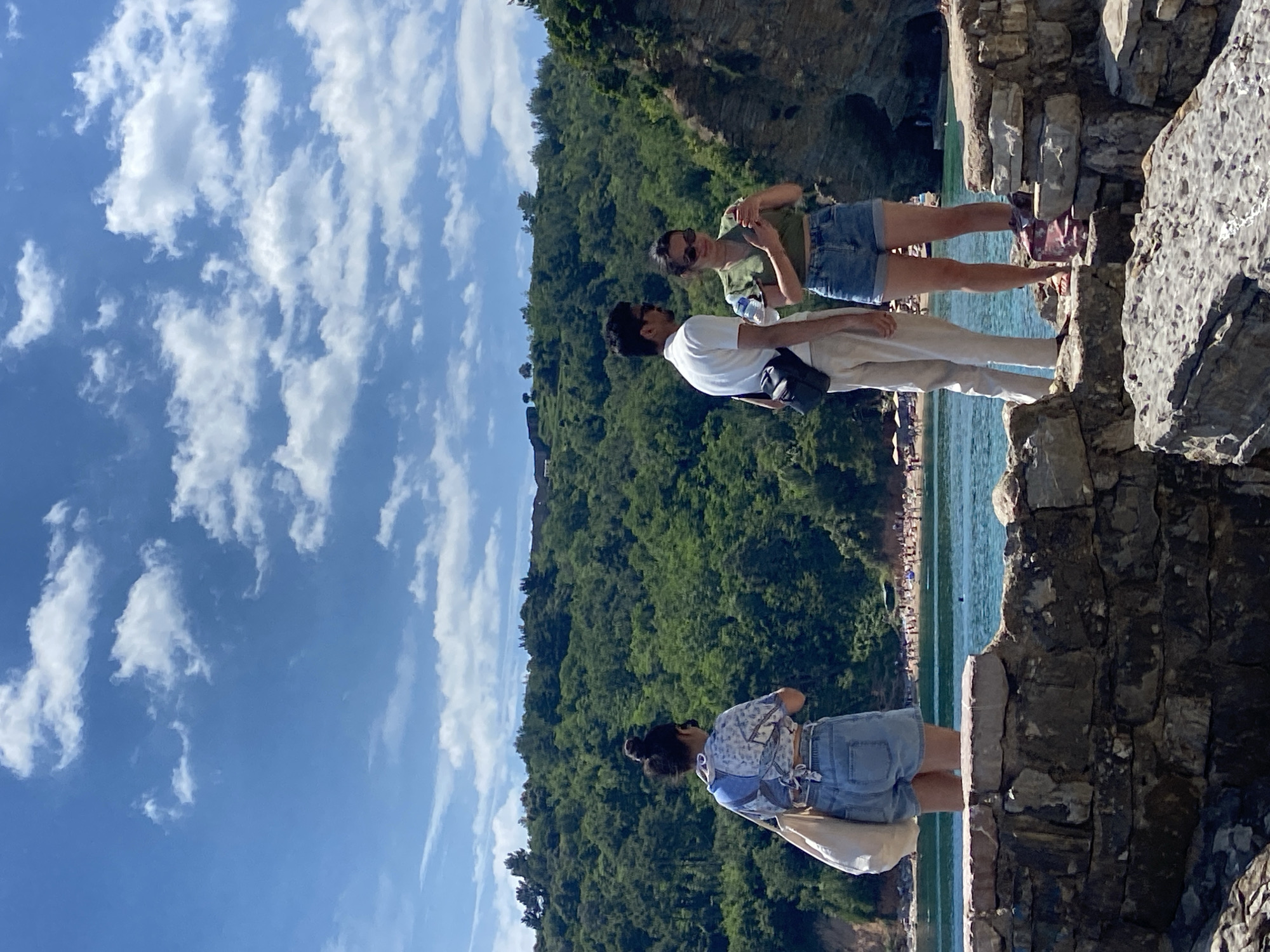 Three people stand on a rocky overlook in Budva, Montenegro, overlooking a beach area. The sky is bright blue with scattered white clouds. The hillside is densely forested. The person to the left is wearing a light blue top and denim shorts, with a white bag. A man in a white shirt and khaki pants stands in the middle, and a woman in a green shirt and denim shorts stands to the right. The rocky area is composed of large stones and provides a clear view of the beach. The image is oriented sideways.