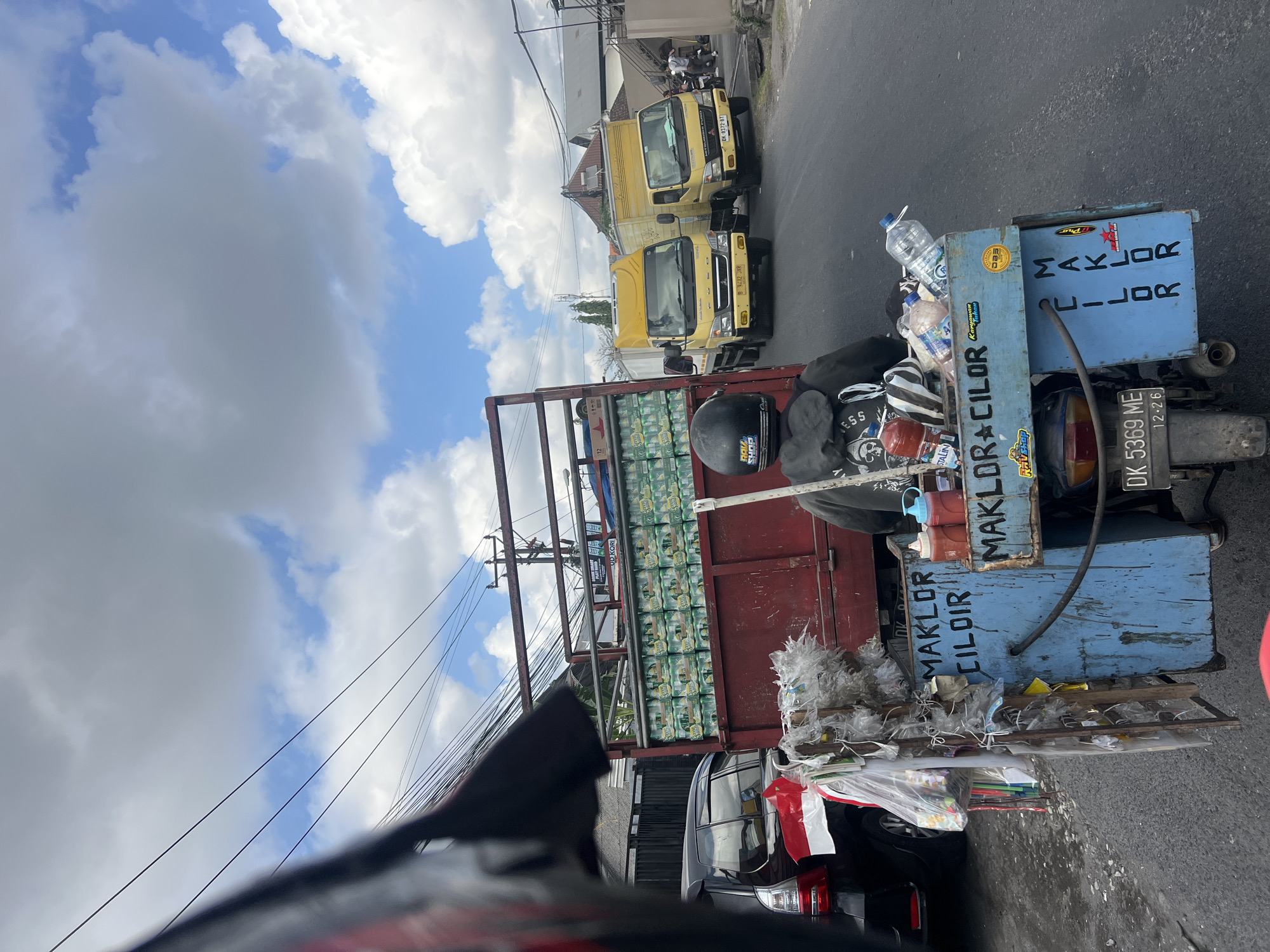 In Denpasar, Indonesia, a street scene unfolds with a focus on a motorcycle carrying a makeshift vendor cart. The cart is laden with bottles of water and what appear to be condiments, such as ketchup and chili sauce, suggesting a food or drink vendor. The motorcycle is a blue, open-back scooter, modified with a blue wooden box structure on the rear. Text on the box reads "MAKLOR CILOR" and "Karyawan Tukang". Attached to the side of the cart is a collection of plastic bags filled with various items, possibly merchandise or supplies.
Behind the motorcycle, a red flatbed truck is parked. The truck's bed is filled with rows of green cans, labeled "GREEN SAND," and stacked neatly. A black SUV is visible to the left of the red truck. In the background, two yellow trucks, a smaller and a slightly larger one, are parked on the right side of the road. The larger yellow truck has the license plate "DK 8372 BT." The scene is set on a paved road under a bright sky with scattered clouds. Utility poles and power lines are visible in the background, typical of urban Indonesian streetscapes. The overall impression is a snapshot of daily commerce on a bustling street in Denpasar.