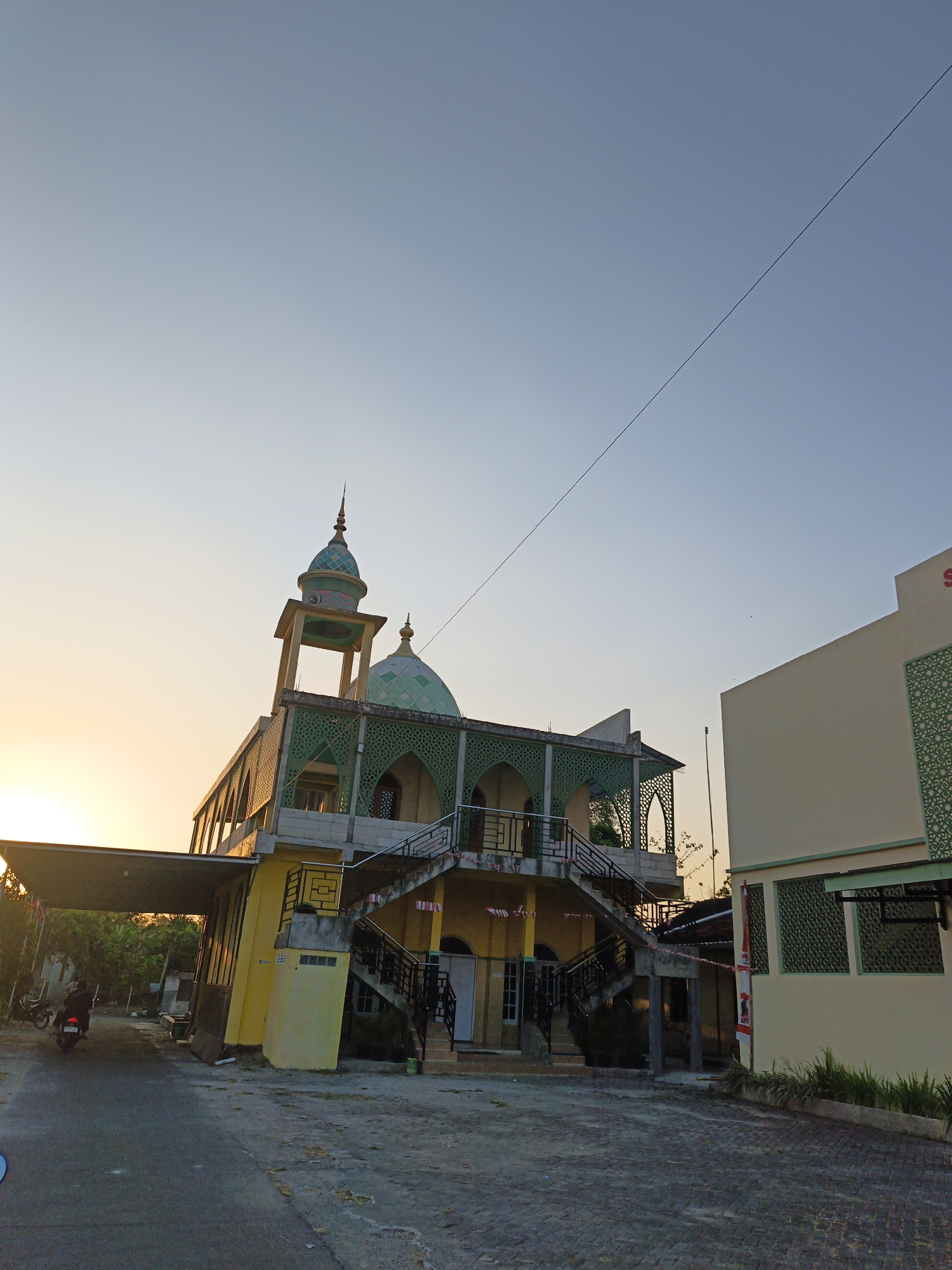 The image depicts the exterior of a mosque in Trucuk, Indonesia, during what appears to be either sunrise or sunset. The mosque has a distinctive yellow facade with green decorative elements and a green dome topped with a spire. An external staircase with black railings leads to an elevated section of the mosque. To the right of the mosque, a pale yellow building with green decorative window panels is visible. The foreground shows a paved area with a road and some vegetation. A motorcycle with a rider is seen on the road to the left, approaching the mosque. The sky is clear and a pale blue, with the warm glow of the sun low on the horizon.