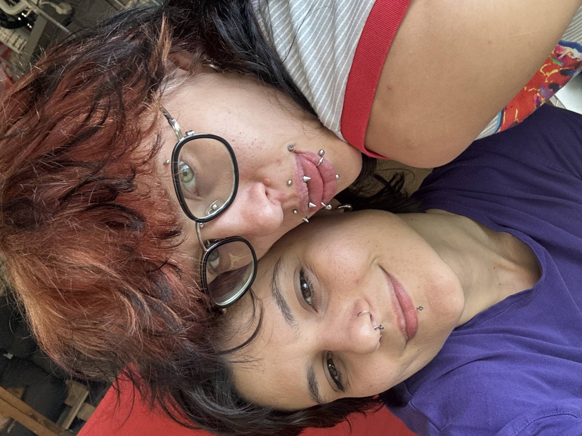 Two individuals are closely positioned for a selfie. The person on the left has red and black hair, wears round glasses, and has multiple piercings around their mouth and tongue. The person on the right, with dark hair, smiles at the camera, showing piercings on their nose and chin. The background is blurred, and the exact location is not determinable from the image alone, therefore the city of Karlukovo, Bulgaria cannot be confirmed.