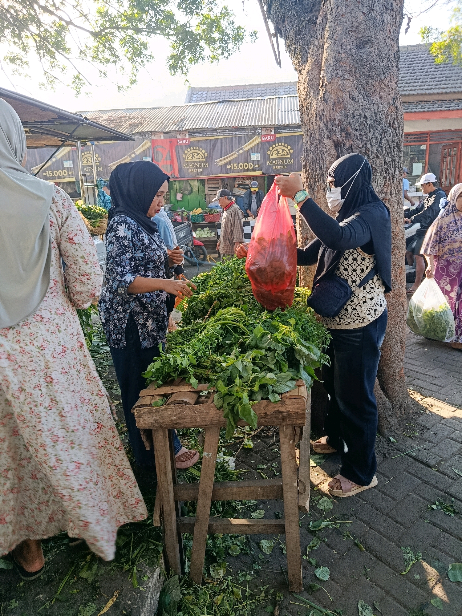 In Bululawang, Indonesia, an outdoor market scene unfolds under dappled sunlight filtering through a large tree. Two women are engaged in a transaction at a stall overflowing with green vegetables. One woman, wearing a black hijab and a patterned blue and white top, is facing away from the camera, her hands near the vegetables. The other woman, also in a black hijab and a white crocheted vest over a dark top, holds a large, translucent red plastic bag filled with red produce. She wears a mask and has a small dark bag slung across her body. A third woman, partially visible on the left, wears a light-colored, floral patterned dress and a light grey hijab. In the background, a stall with awnings displays promotional banners and what appear to be cigarettes. Further back, other market-goers and stalls are visible, with a motorcycle parked near one stall. The ground is paved with dark interlocking bricks, scattered with green leaves and debris, suggesting the dynamic nature of a bustling market.