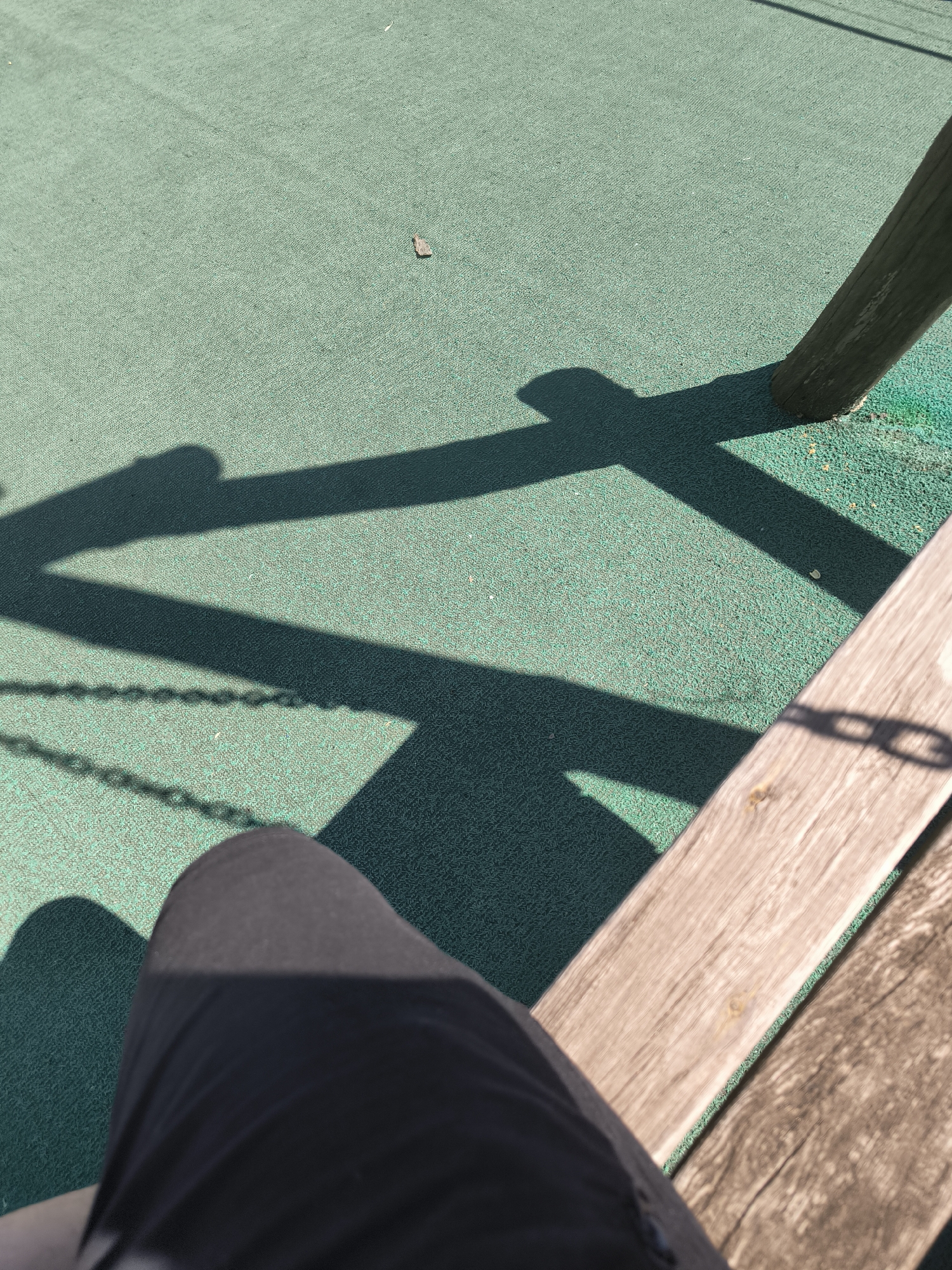 The image shows a person's legs and a shadow on a green surface. The shadow is cast by a structure that appears to be a swing set, with chains visible. A wooden post is also visible, casting a shadow. The green surface appears to be a rubberized playground surface. The scene is likely outdoors, possibly in a park or playground in Chișinău, Moldova. There are no other people or animals visible in the image.