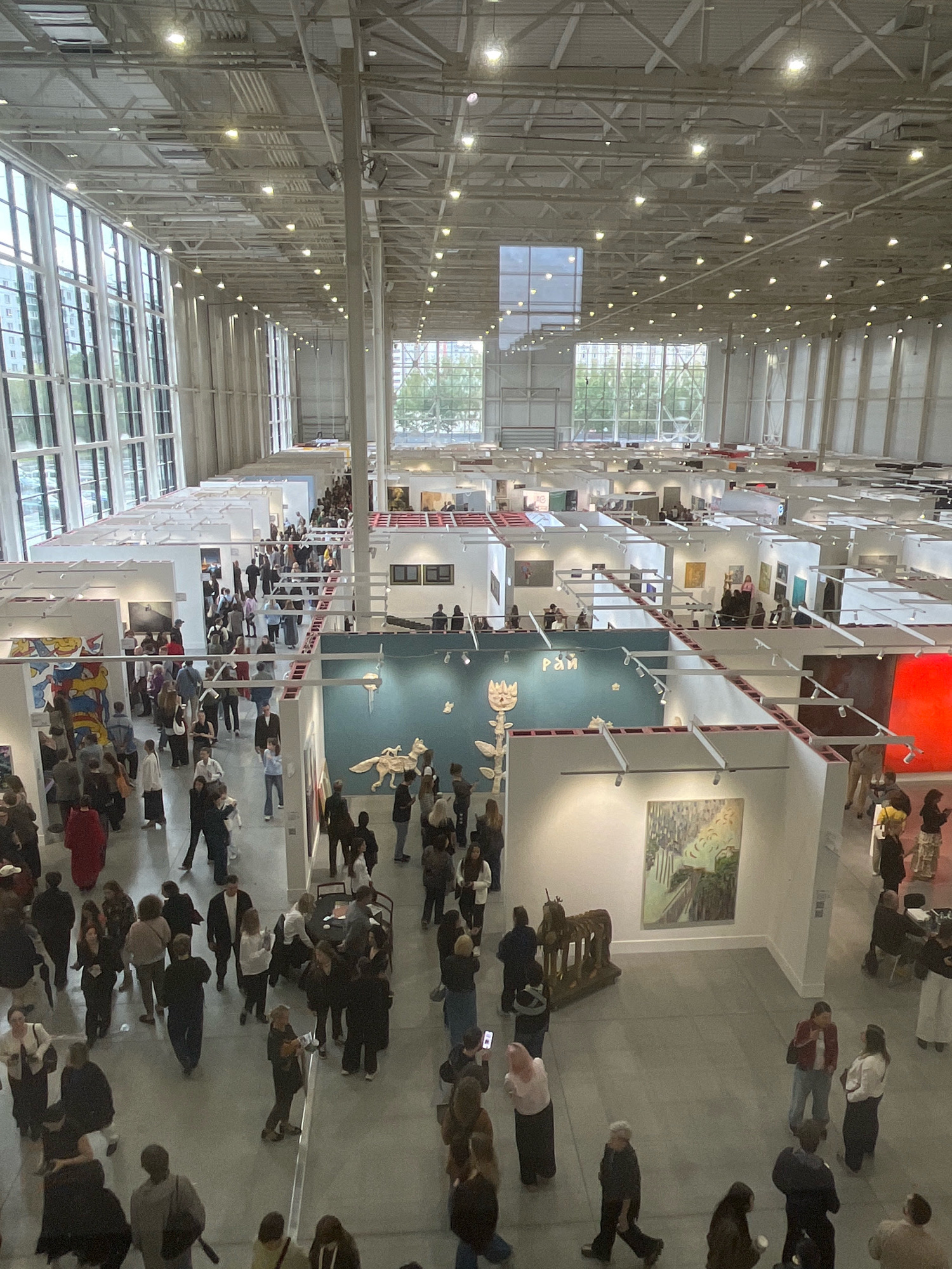 The image depicts a large indoor art exhibition or fair within a spacious, modern exhibition hall in Khimki, Russia. The hall features high ceilings with exposed industrial-style lighting and a grid of metal beams. Large windows along one side of the hall allow natural light to enter. The exhibition space is divided into numerous white-walled booths displaying various artworks, including paintings and sculptures. A significant number of people are present, milling around the booths, observing the art, and interacting with each other. They are dressed in casual attire. The overall atmosphere appears to be that of a busy public event, possibly a weekend art fair or gallery opening.
