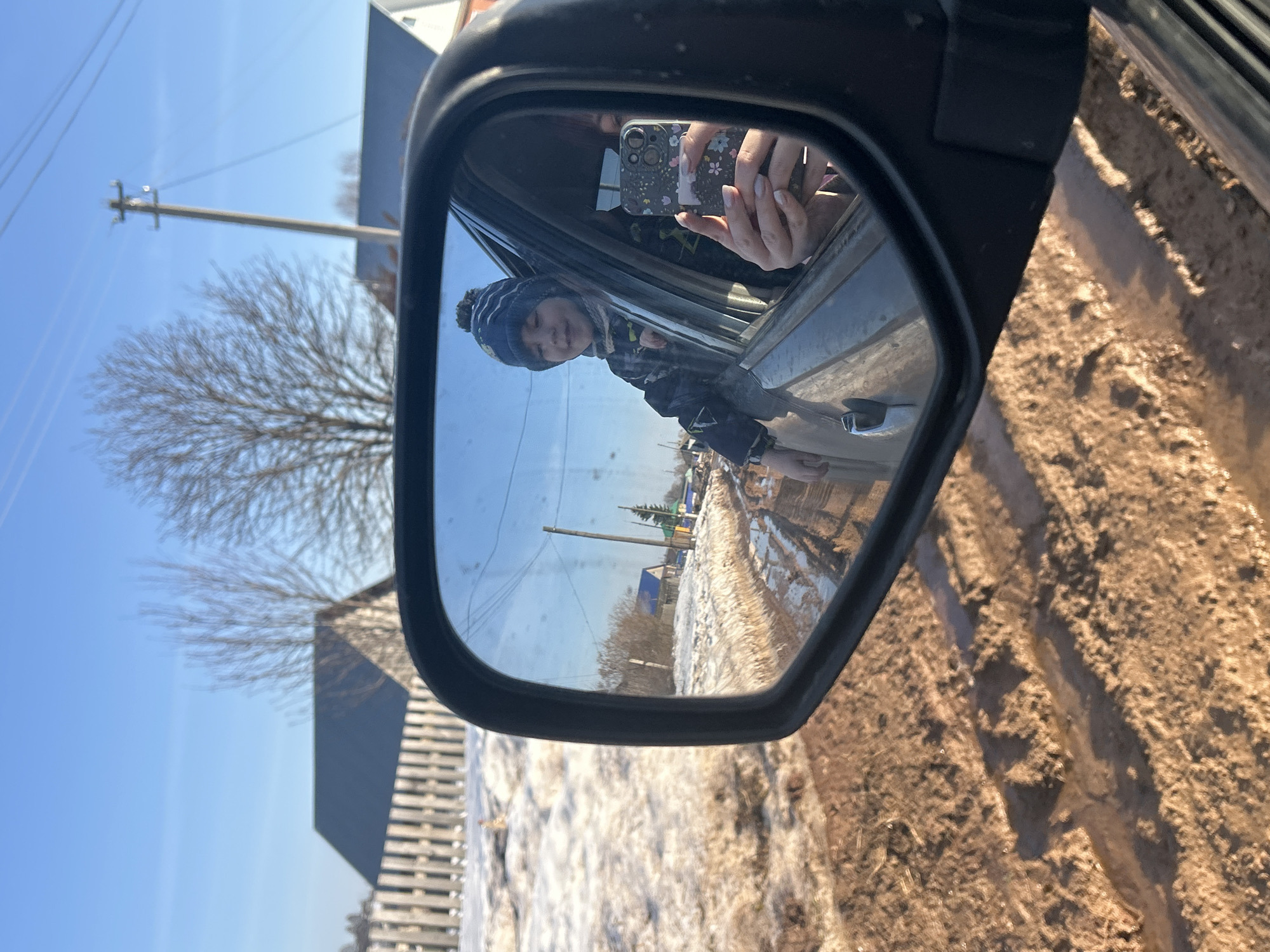 A side view mirror captures a reflection of a person in a blue winter hat, partially visible out of a car window. In the foreground of the mirror's reflection, a snow-covered road stretches along a rural area. The surrounding environment features bare trees and utility poles under a clear blue sky. The foreground outside of the mirror shows a dirt road. The phone is an Apple iPhone. The mirror is black plastic. The photo appears to be taken near Фоки, Russia.