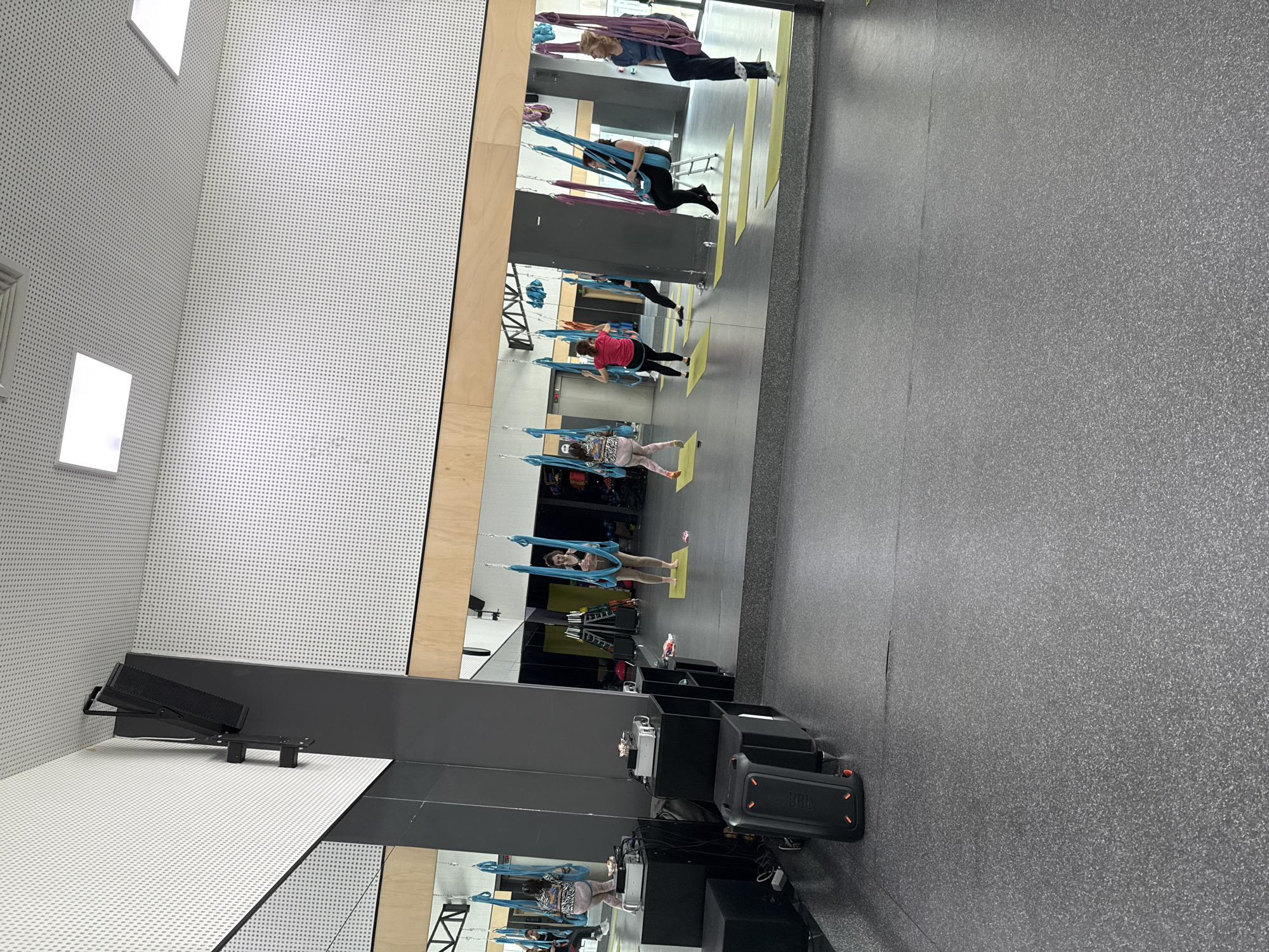 Here is a description of the image:
The image shows an indoor space, possibly a fitness studio, where people are engaging in aerial yoga. The room has a wall with a large mirror that reflects the activities taking place. Individuals are suspended in yoga hammocks or silks, performing various poses. The room's decor includes a white, dotted wall pattern and wooden accents. There's fitness equipment visible, suggesting a professional or dedicated space for such activities. The floor is a consistent, neutral gray color. The overall lighting is bright and even, illuminating the scene clearly. The location is Unknown, Unknown.