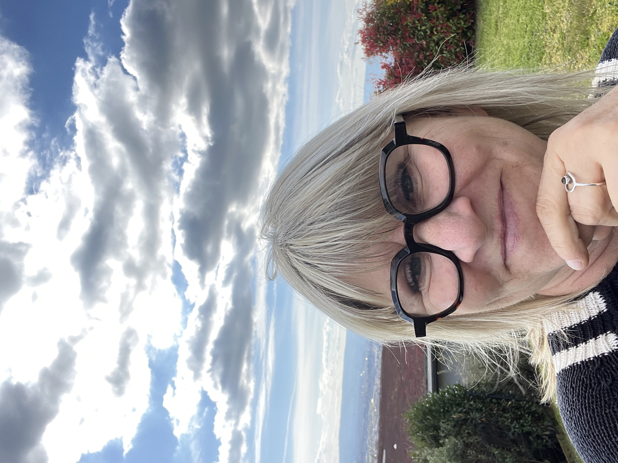 A selfie shot outdoors shows a middle-aged woman with short, gray-blonde hair wearing black-framed glasses and a black-and-white striped sweater. The background features a cloudy blue sky, a building or landscape with red tones, and greenery, possibly bushes or trees. A silver ring with a small stone is visible on her finger. Based on the prompt information, this image was taken in Chambœuf, France.