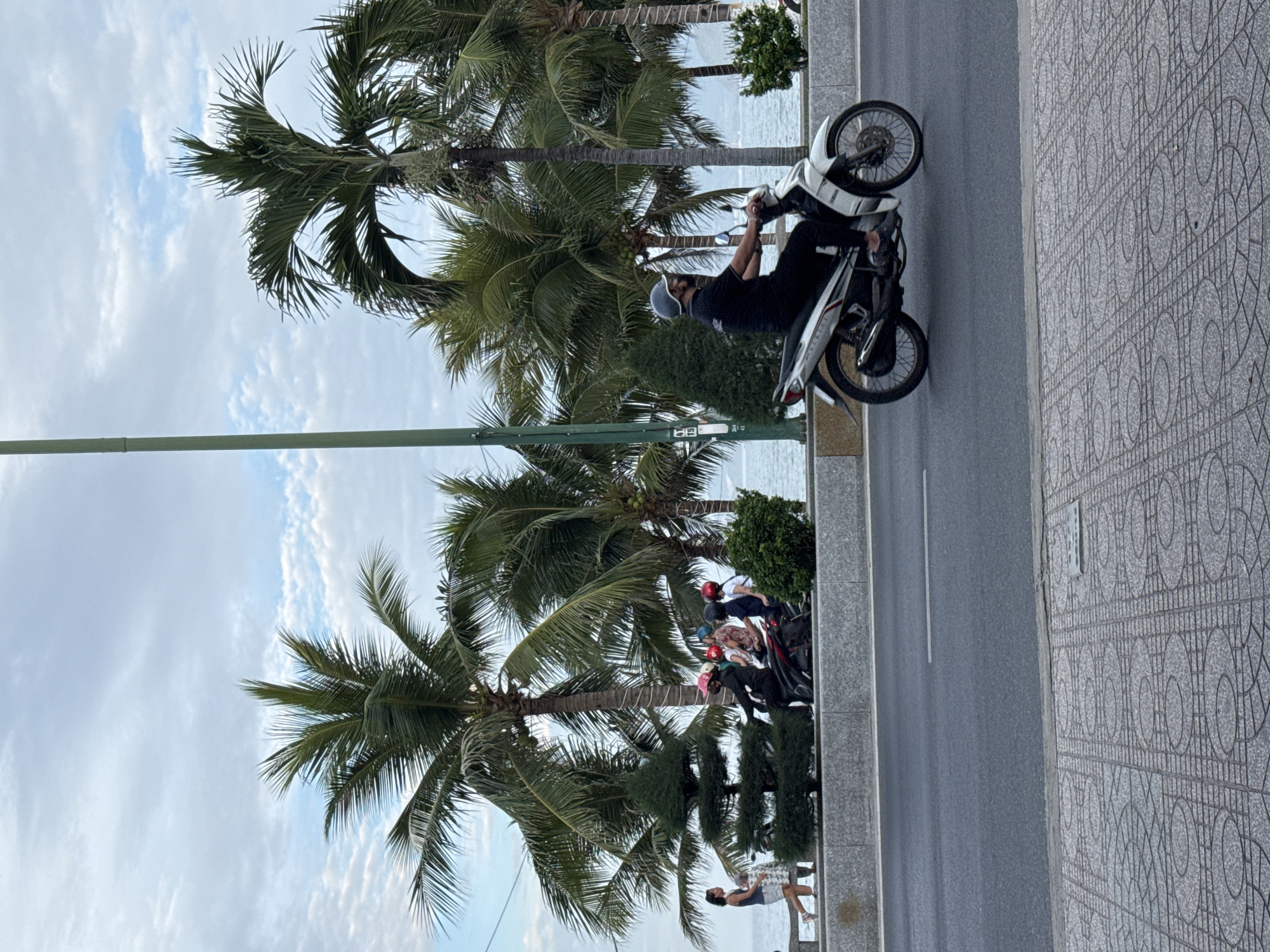 A man wearing a dark shirt, dark pants, a light-colored helmet, and a black face mask rides a white and black motorcycle along a paved road in Nha Trang, Vietnam. The road is bordered by a sidewalk with a decorative patterned surface on the right and a raised concrete barrier on the left. Beyond the barrier, palm trees and other greenery line the street, with the ocean visible in the background. Several other motorcycles and people are visible on the sidewalk area, including a woman in a dark tank top and shorts walking away from the camera, and a group of people on motorcycles wearing helmets. The sky is overcast with clouds.