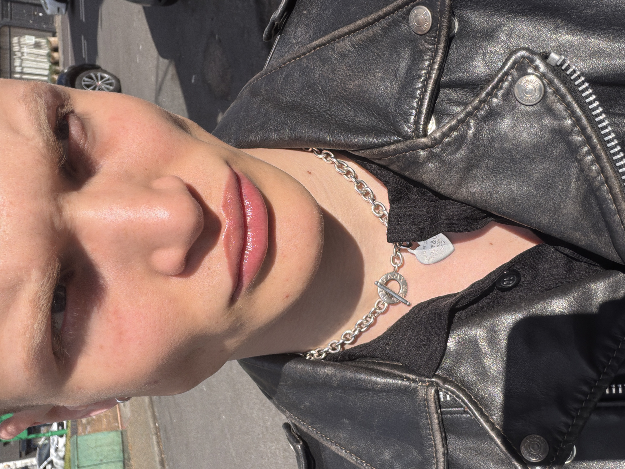 The image is a close-up, slightly angled view of a person wearing a black leather jacket and a silver chain necklace with a toggle clasp. The person's face is visible, featuring closed eyes, fair skin, and lips with a glossy finish. The leather jacket has visible stitching and metal buttons with the word "SCHOTT" inscribed on them. A black garment is visible underneath the jacket. Part of a street with a parked car is visible in the background. Based on the branding on the necklace ("Tiffany & Co. New York 925"), the location could possibly be in or near a Tiffany's store, which may be located within London.