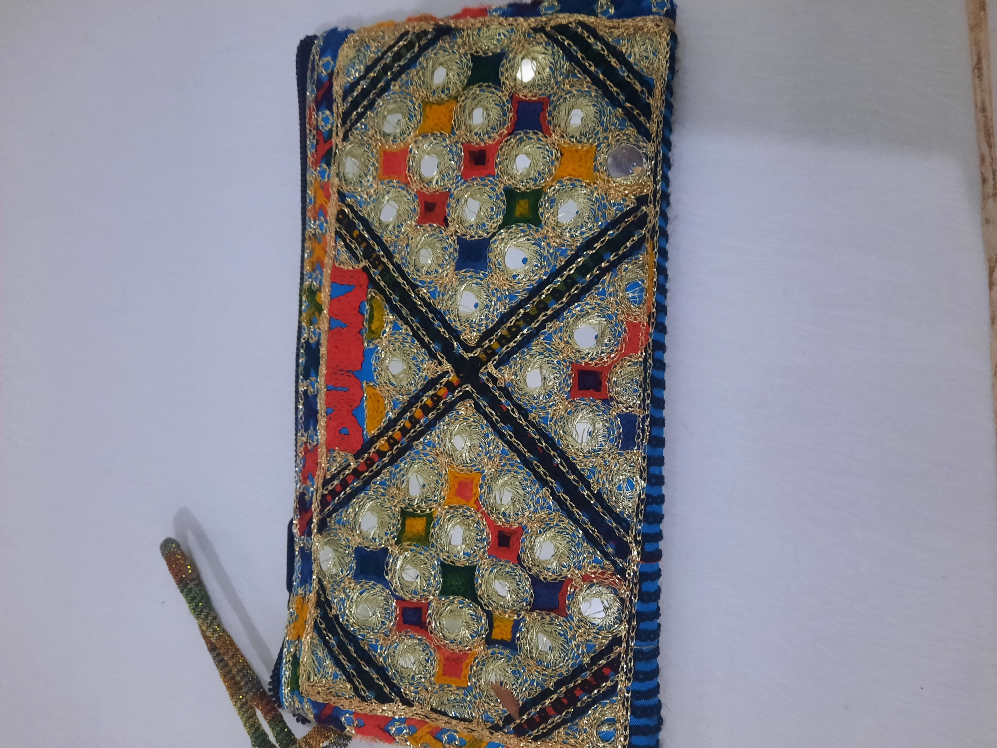 A colorful embroidered clutch with mirrors is displayed on a white surface. The clutch features a geometric pattern of squares and circles, with intricate gold thread embroidery and small mirrored embellishments. The background appears to be a plain white surface, likely a table or floor. There are no people, animals, or identifiable city landmarks present in the image. The location in Lhokseumawe, Indonesia, is not visually represented in this photograph.