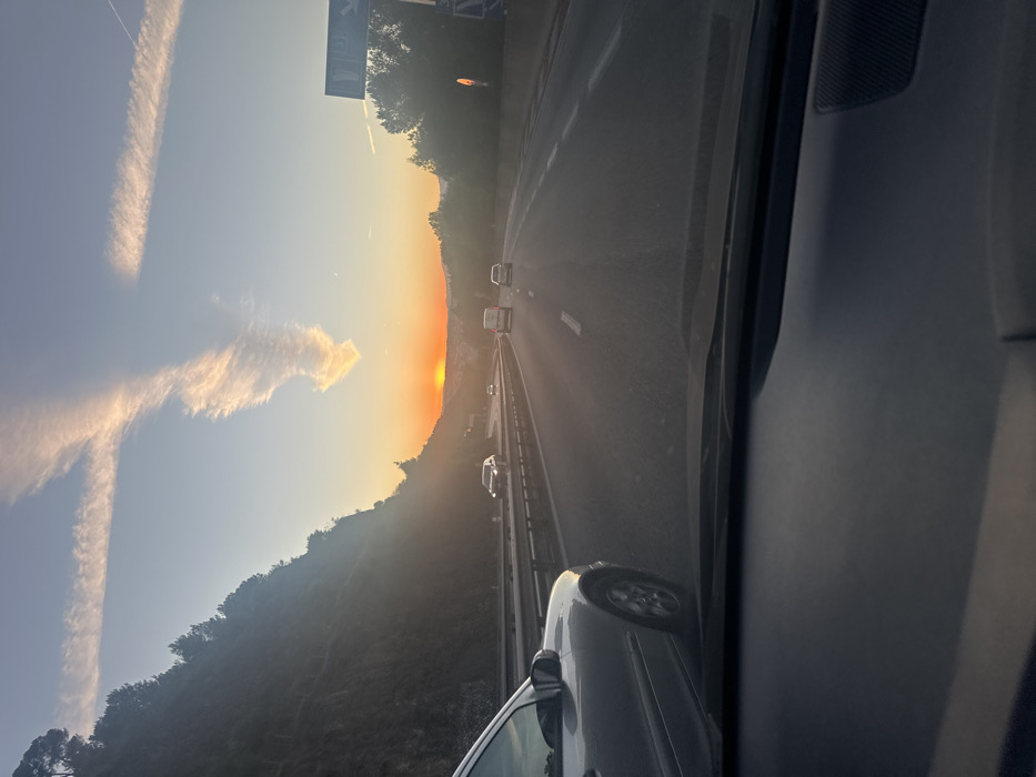 The image depicts a multi-lane highway scene at sunset, viewed from inside a vehicle looking forward through the windshield. The location is consistent with the mountainous terrain surrounding La Trinité, France, likely on the A8 motorway due to the steep, tree-covered hillside on the left.
The setting sun, a bright orange disc, is visible low on the horizon, slightly to the left of the road's center, casting strong glare and long shadows. The sky transitions from light blue at the top to a warm orange and yellow glow near the horizon. Two prominent, elongated contrails or cirrus clouds form an "X" shape high in the sky, with one arm extending downwards and broadening.
The highway features at least two lanes of traffic moving away from the viewer. White lane markings are visible on the asphalt. A metallic crash barrier runs along the left side of the road, separating it from the steep, dark green, tree-covered hillside. On the right, further trees and a partially obscured blue road sign with white text or symbols are present.
Several vehicles are on the road, traveling in the same direction. A silver sedan is partially visible in the left foreground, adjacent to the viewpoint vehicle. Further ahead, a dark sedan is in the left lane, followed by a white van or SUV in the right lane, and another light-colored sedan further ahead in the right lane. The dashboard of the viewpoint vehicle, dark and featureless, occupies the lower portion of the frame. No individuals are explicitly visible.