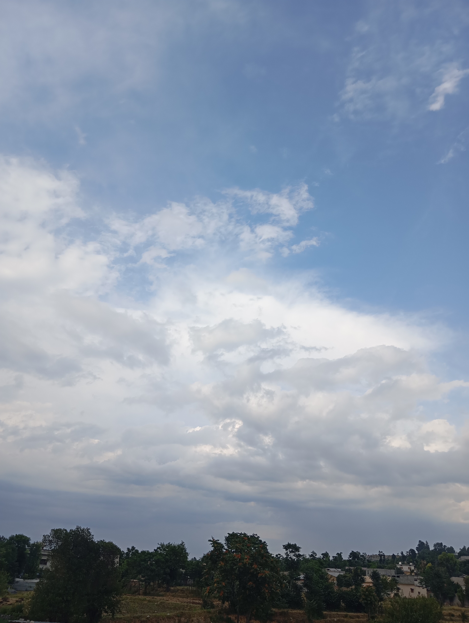 The image depicts a sky filled with a mix of white and grey clouds, with patches of clear blue sky visible towards the top. Below the sky, a treeline with various types of trees, some appearing lush green and others with darker foliage, forms the horizon. In the distance, there are glimpses of buildings and rooftops, suggesting a semi-urban or rural setting. The ground in the foreground appears to be a mix of dry earth and vegetation. The lighting suggests it is daytime, possibly late afternoon or early morning, given the diffused light and the varied cloud cover. There are no people or animals visible in the image. The overall scene is tranquil and natural, with a hint of human habitation in the background. The location is identified as Dangari, Pakistan.
