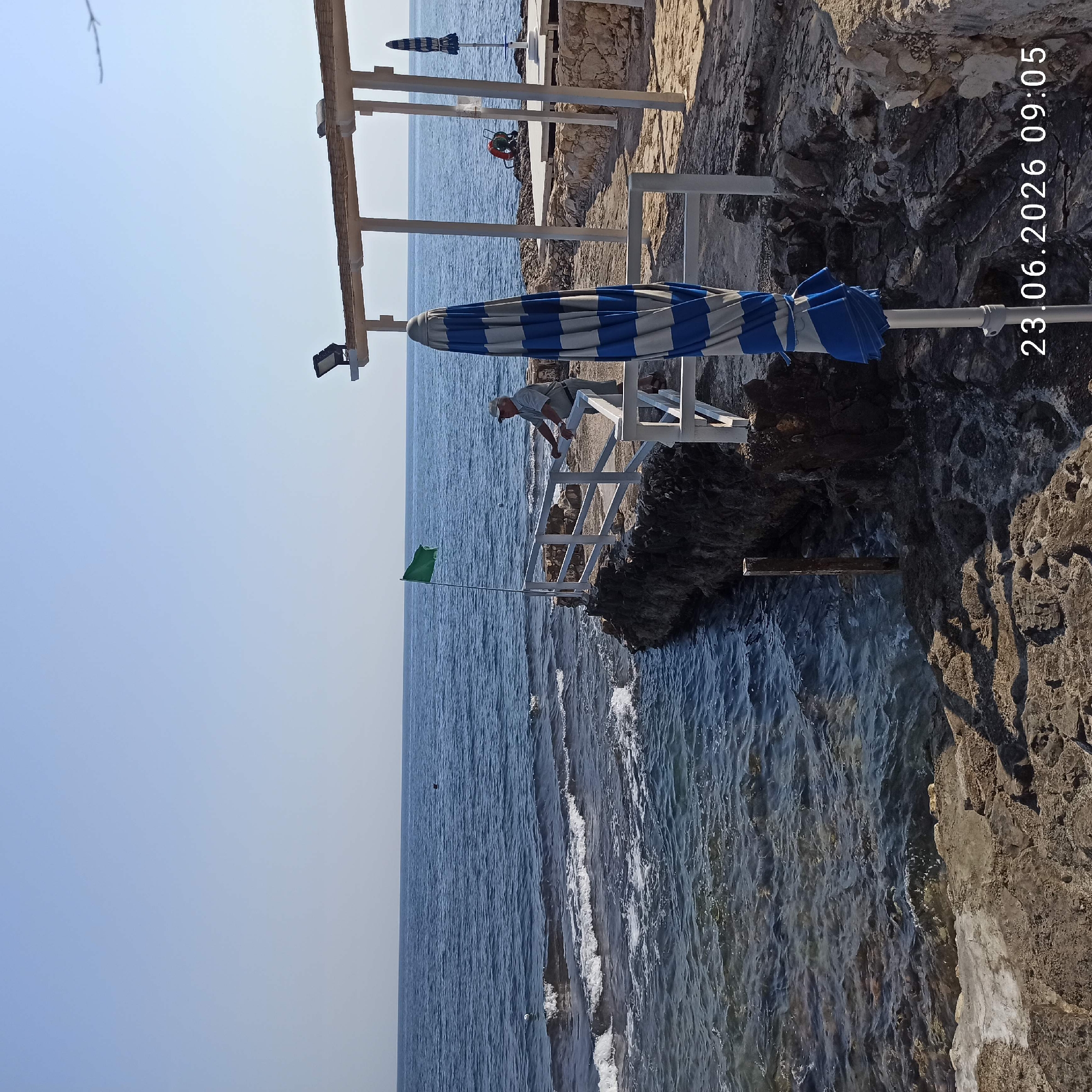 The image depicts a rocky coastline in Molfetta, Italy, with the Adriatic Sea on the left. A white railing runs along the edge of the rocky outcrop, with a man in a light-colored shirt and cap leaning on it, facing the sea. To the left of the man, a green flag flies from a pole. In the foreground, a partially closed blue and white striped umbrella stands upright. In the background, a shaded structure with white posts and a thatched roof is visible, along with another umbrella. A life preserver is also visible near the structure. The water is calm with small waves breaking on the rocks. The sky is clear and blue. A timestamp "23.06.2026 09:05" is overlaid on the bottom right corner of the image.