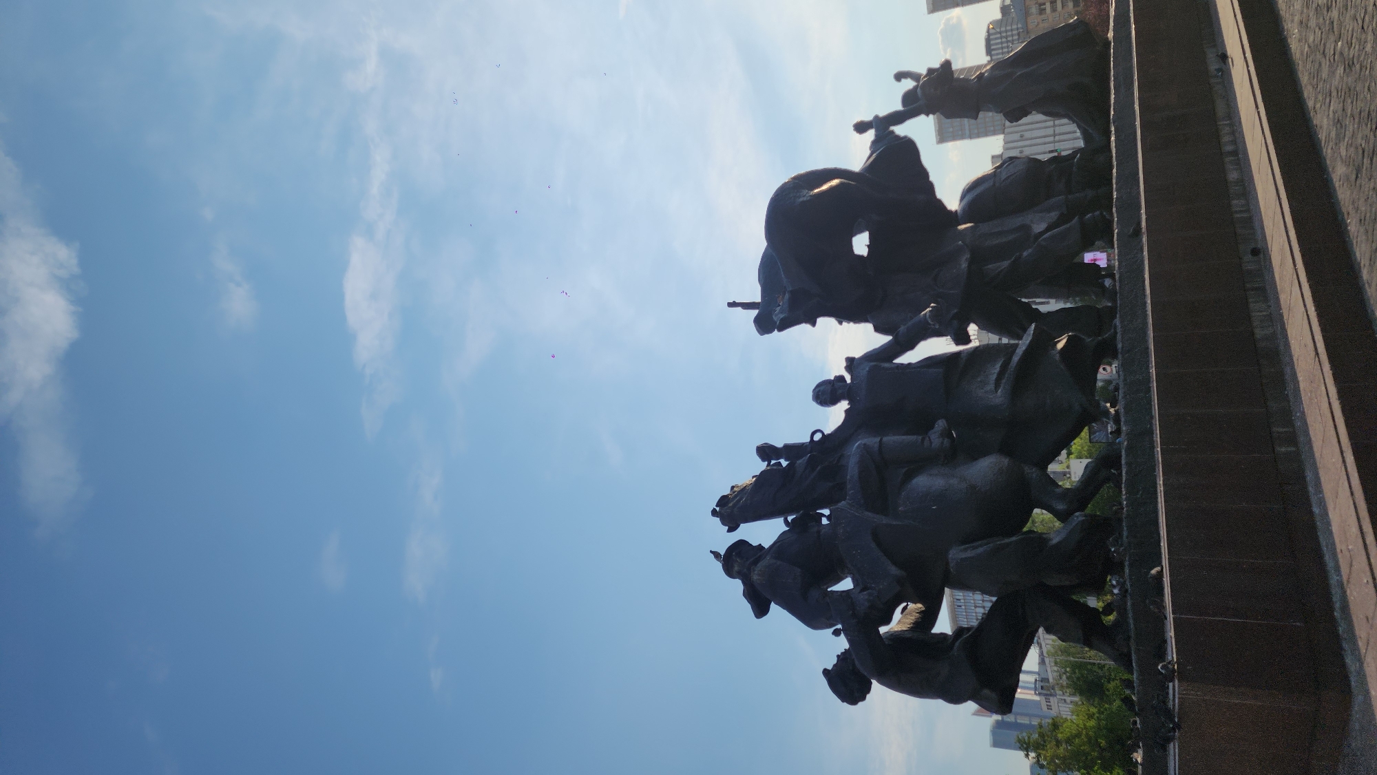 The image shows a monument in Moscow, Russia, depicting figures on horseback. The dark bronze monument shows multiple people and horses in a dynamic, forward-moving action. The monument is situated outdoors, with a blue sky with wisps of clouds as a backdrop. A stone base is visible under the monument, and buildings are noticeable in the distance, hinting at an urban setting.