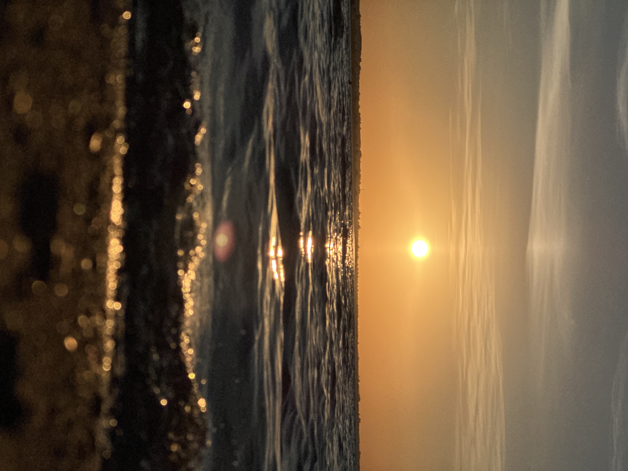 The image depicts a sunset over a body of water, possibly a lake or sea. The sun, a bright orange orb, is low in the sky, casting a warm glow and creating a shimmering reflection on the water's surface. The sky is a gradient of soft oranges and blues, with wispy clouds stretching horizontally. In the distance, a faint silhouette of a coastline with some vegetation is visible. The foreground is dominated by the dark, textured surface of the water, with small waves creating ripples and catching the light. The very bottom of the image is out of focus, showing a blurry brown surface, likely a beach or the ground. The scene conveys a sense of tranquility and natural beauty. There is no visible indication of the city of Kokshetau, Kazakhstan in this image.