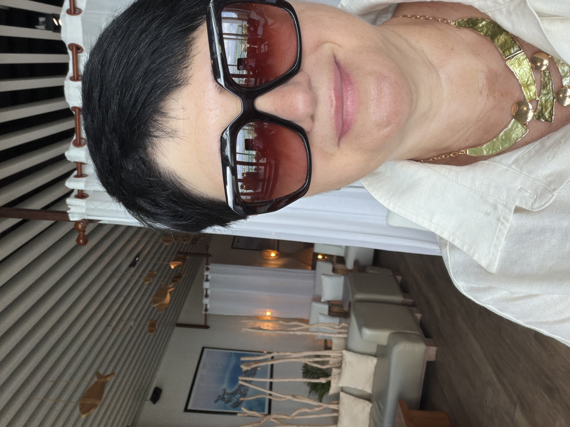 A woman with short black hair and wearing large, dark-rimmed sunglasses and a white collared shirt poses for a selfie. She is wearing a chunky gold necklace with green accents. The background suggests an indoor dining or lounge area, with light-colored furniture and wooden flooring. Suspended wooden fish decorations are visible on the ceiling. Curtains hang in the background, and framed artwork adorns the wall. The overall ambiance suggests a tropical resort or upscale establishment, potentially located in the city of Malé, Maldives, given the light and airy decor often associated with such locations. The sunglasses reflect a scene of the interior and possibly an outdoor view.
