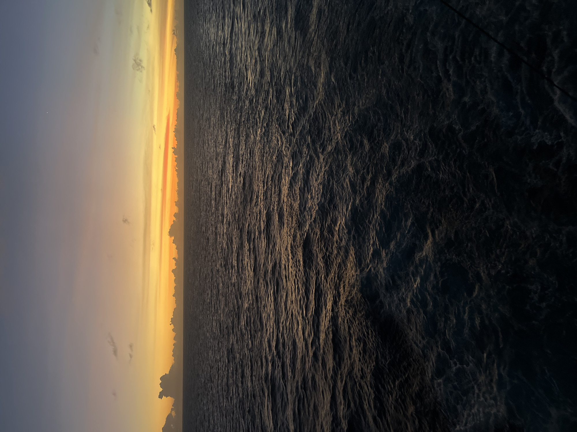 The image depicts an ocean at sunset. The left side of the image shows the sky transitioning from blue to orange and yellow hues, with some clouds visible near the horizon. The right side of the image shows the ocean, with waves and textures visible in the water. The water appears dark, reflecting the fading light of the sunset. The location is Unknown, Unknown.