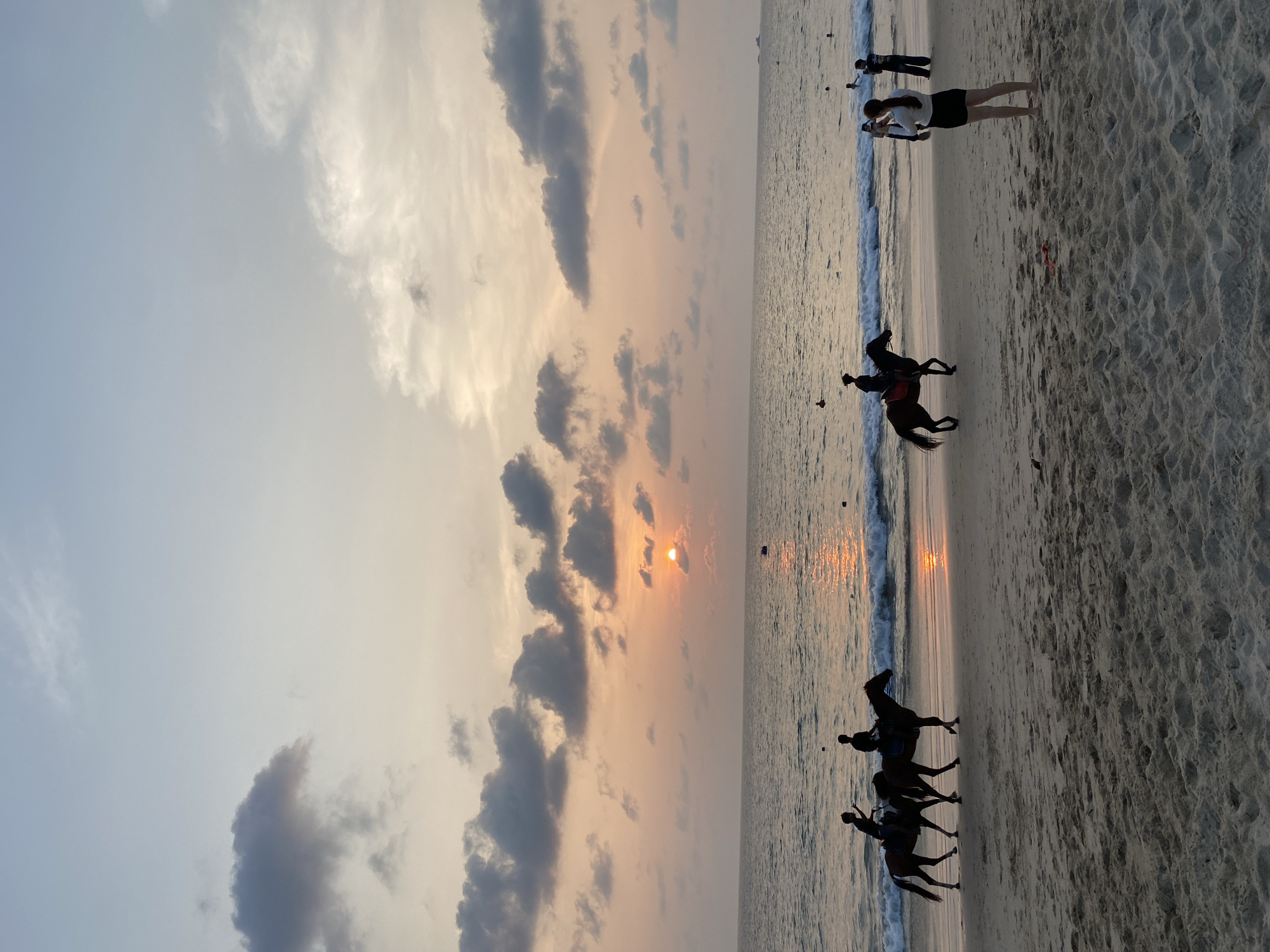 The image captures a vibrant sunset scene on a beach in Choeng Thale, Thailand. The sky, occupying the upper portion, is illuminated by the setting sun, visible as a bright orb low on the horizon. Warm hues of orange, yellow, and pink blend with cooler greys and blues, interspersed with dark, dramatic clouds.
Below the sky, the ocean reflects the sunset's golden glow, with gentle waves breaking onto the sandy shore. The beach itself stretches across the foreground, displaying textured sand.
Several subjects are engaged in evening activities. On the left side, three horses, each with a rider, are silhouetted as they wade through the shallow water near the shoreline. Their dark forms create a striking contrast against the bright reflections on the water.
On the right side of the image, several people are visible on the beach and in the water. A person, possibly a woman, stands prominently on the sand in the mid-ground, facing the ocean. Further down the beach and into the water, other figures, including what appear to be two children playing near the water's edge, can be seen enjoying the serene atmosphere.
The time of day is clearly sunset, as indicated by the sun's position and the golden light illuminating the scene. The weather appears partly cloudy. The overall mood is peaceful and leisurely, characteristic of a tropical beach evening. There is no visible text in the image.