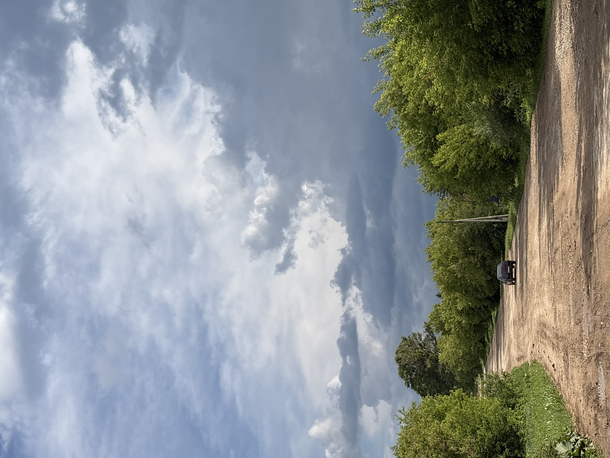 A dark-colored sedan is traveling away from the camera on a dirt road, which curves to the left. The road is bordered by lush green trees on both sides, with a few taller trees visible in the background. The sky above is filled with dramatic, heavy grey clouds with some patches of lighter sky peeking through. The scene appears to be in a rural or semi-rural area, possibly on the outskirts of the village of Kumyony, Russia, given the characteristic dirt road and vegetation. The lighting suggests a cloudy day, possibly before or after a rain shower.