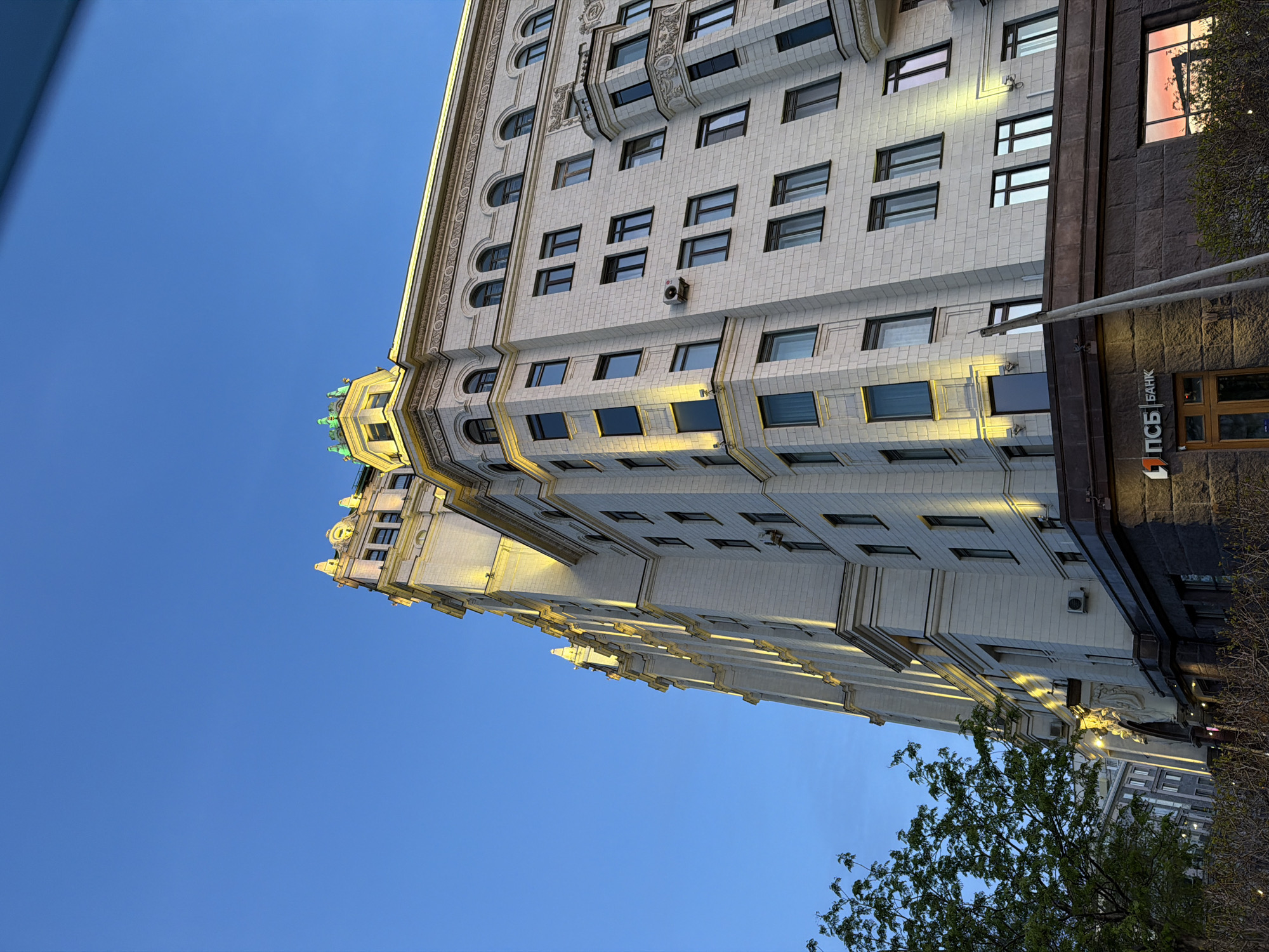 The image depicts the Kudrinskaya Square Building in Moscow, Russia, taken from a low angle pointing upwards. The architecture is Stalinist neoclassical, showcasing numerous windows, ornate details, and setbacks as the building ascends. The lower portion of the building contains a PSB (Promsvyazbank) bank sign. The sky is a clear blue. A tree branch is visible in the foreground.