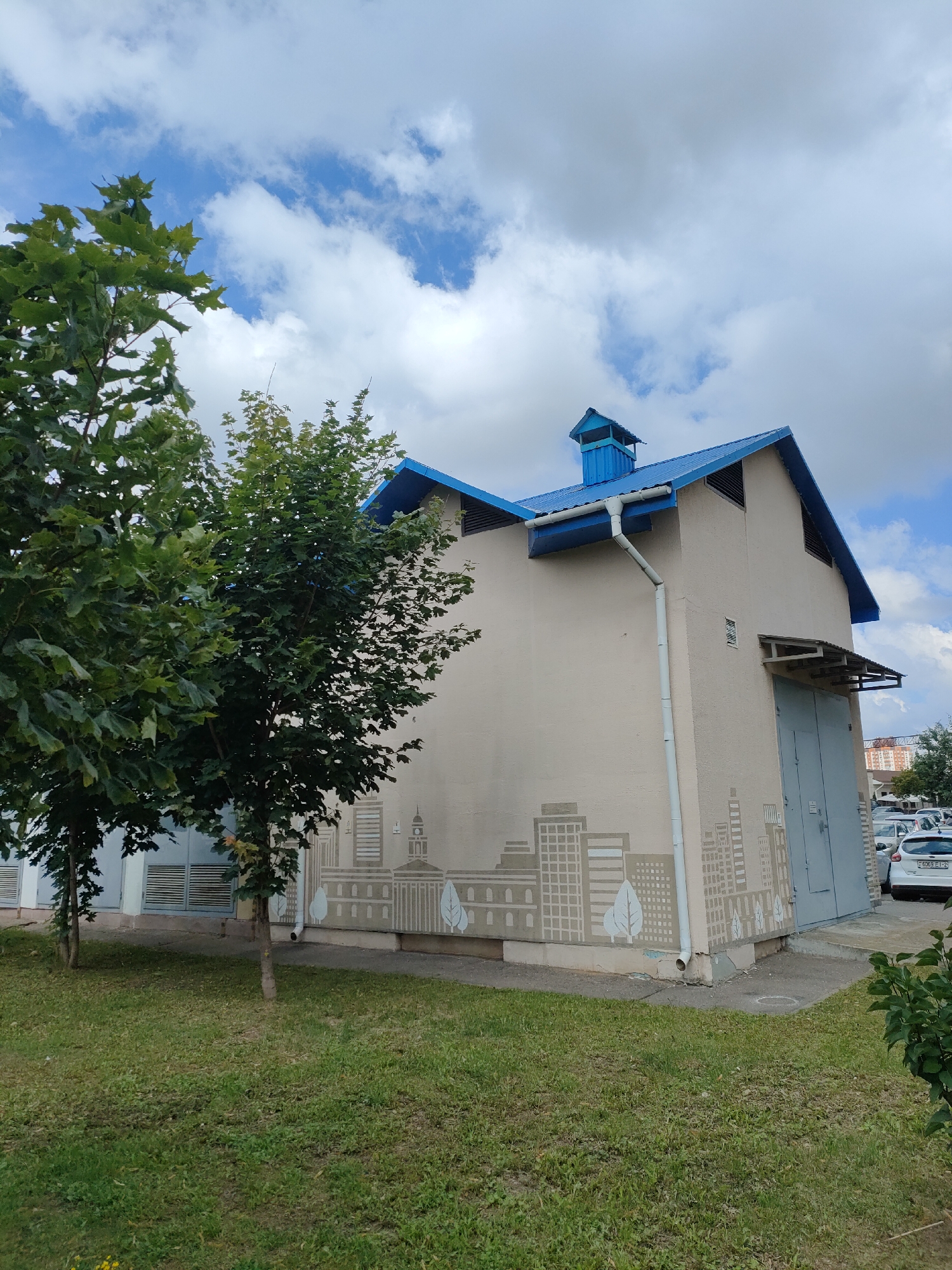 This is a full-shot, eye-level, outdoor, daytime, color photograph of a building with a mural on its side, taken in Minsk, Belarus. The building is light beige with a blue, sloped roof. It has a large, gray, double-door garage entrance on the right side. On the lower part of the building's side wall, there is a mural depicting stylized buildings and trees. To the left of the building, there are two trees with green leaves. A patch of green grass is in the foreground. In the background, there are several parked cars and other buildings, including some with red accents on their roofs. The sky is blue with white clouds.