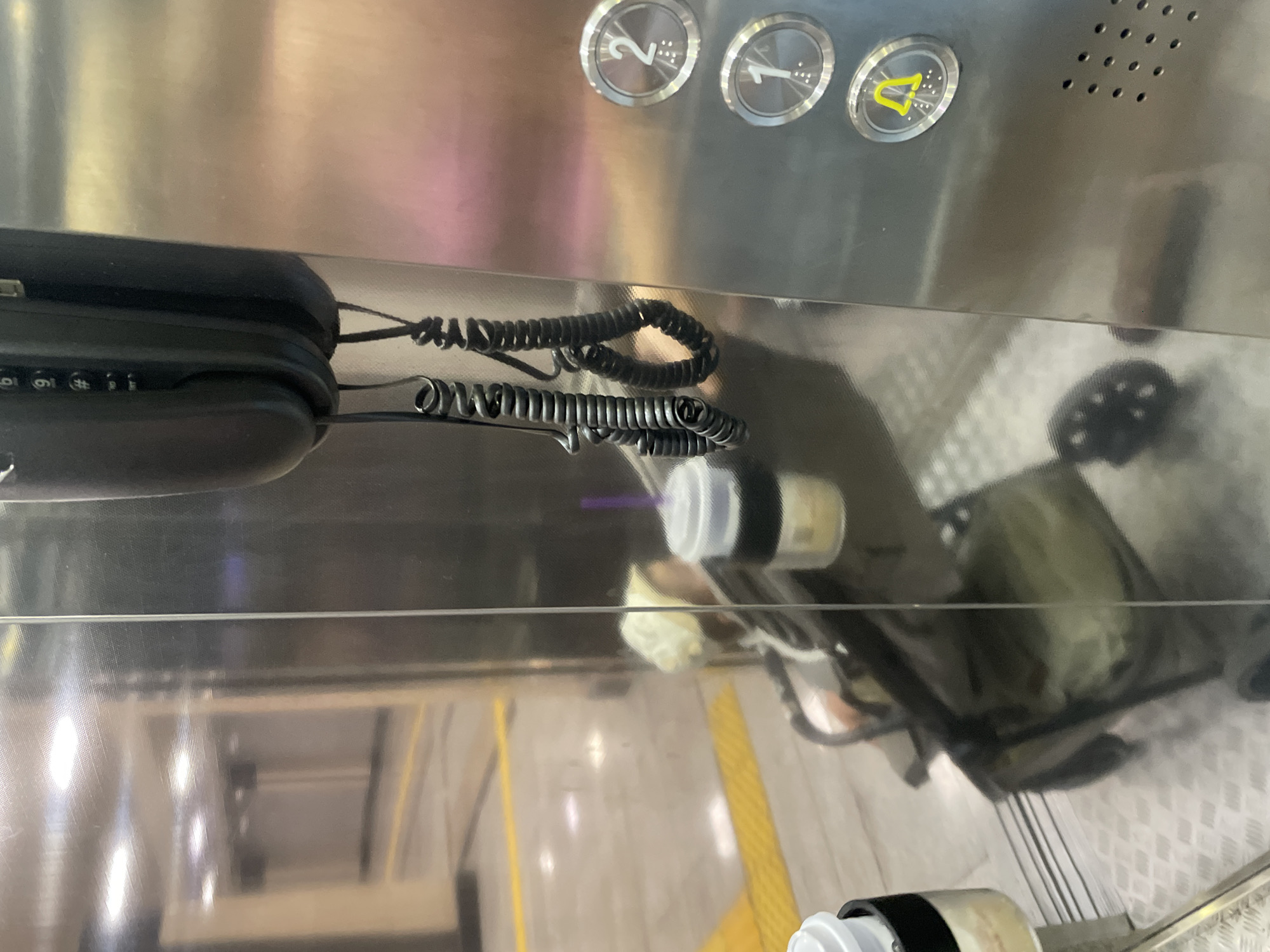 An elevator interior, likely in Almaty, Kazakhstan, features a person seated in a stroller, holding a disposable cup with a purple straw. The elevator walls are made of reflective stainless steel, creating a mirror-like effect that shows a blurred reflection of a hallway with yellow markings on the floor. A black corded telephone is mounted on the wall, with its coiled cords hanging down. Elevator buttons are visible on the right side, including illuminated buttons for floors 1 and 2, and a button with a yellow alarm symbol. The floor of the elevator is a metallic grid pattern.
