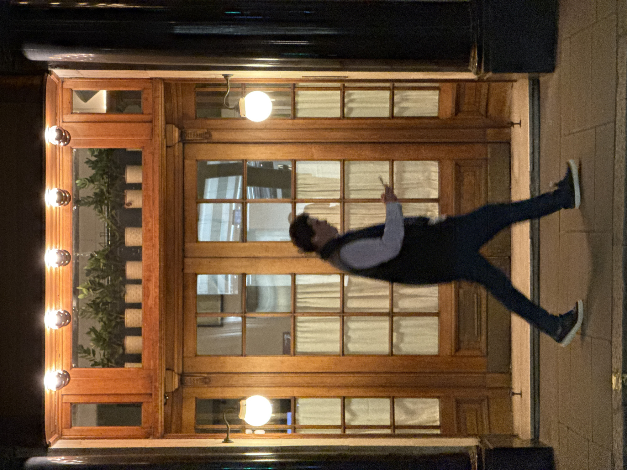 The image captures a person walking past a traditional wooden building entrance at night in what appears to be an urban setting, likely London. The building entrance features double doors and numerous glass panes framed by rich brown wood. On the left side of the entrance, three round, silver-colored sconce lights are mounted vertically, casting a warm glow. Above the right door, a spherical light with a curved arm also illuminates the facade.
Through the windows on the left, several potted green plants are visible on a ledge, adding a touch of greenery to the scene. The interior visible through the windows appears softly lit, with some sections showing light-colored blinds or curtains.
A person with dark hair, wearing a white shirt, a dark vest or jacket, dark trousers, and dark shoes with white soles, is depicted mid-stride. They are holding a smartphone in their right hand, seemingly looking at it or interacting with it. Their movement is captured with noticeable motion blur, particularly on their lower body and the phone, indicating they are in motion.
The time of day is clearly night, as inferred from the dark surroundings and the illuminated architectural features. No legible text is visible in the image.
