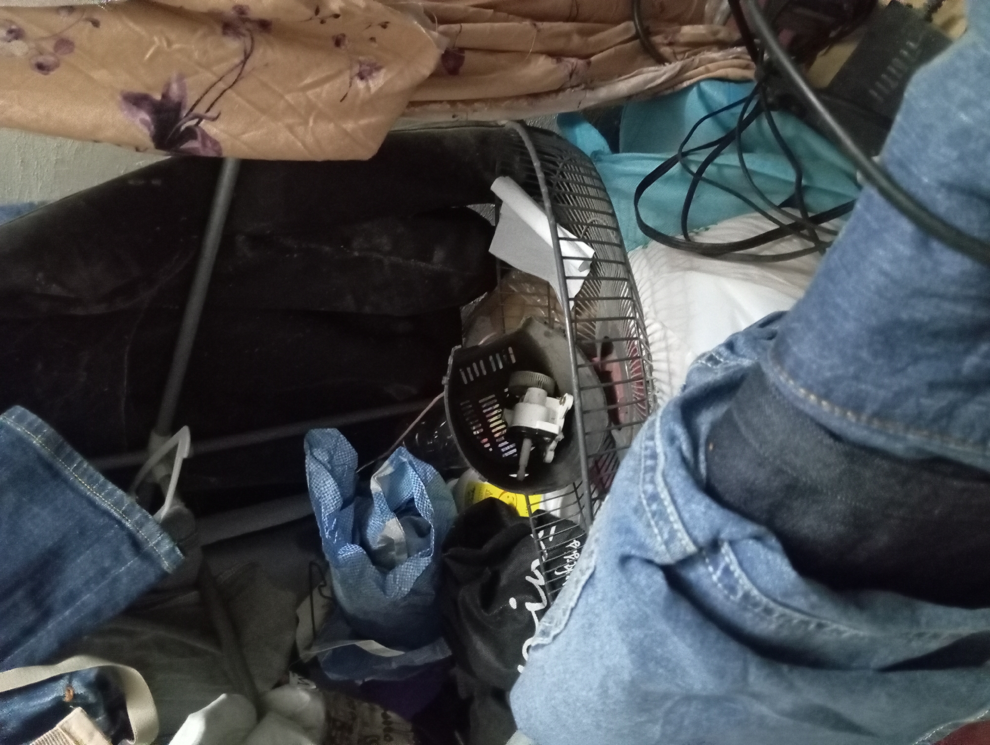 The image depicts a cluttered indoor scene. In the foreground, a portion of blue denim clothing, likely jeans, is prominently displayed. Behind this, a black fabric item, possibly a shirt or jacket, is visible, with white lettering on it that appears to spell "ini" followed by other indistinct characters. To the right and slightly behind, a wire basket contains a small white object with a motor, possibly part of a broken appliance or fan. Behind the basket, there is a blue plastic bag and more black fabric. Further to the right, a beige fabric with a floral pattern hangs, showing shades of brown and purple flowers. The background consists of a textured wall. The scene suggests a storage area or a corner of a room where various items have been gathered. No specific location within Jogosari, Indonesia, can be determined from the visual information.