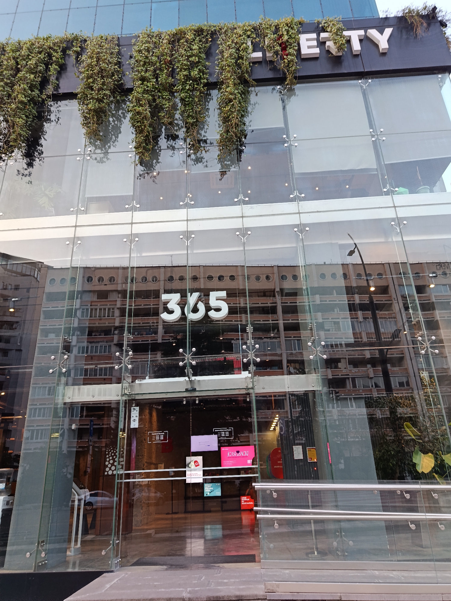 The image shows the exterior of a modern building with a glass facade. The building features a sign at the top that reads "Lerty" in white, block letters, with green vines cascading down from above. Below the sign, the glass facade reflects the interior of the building, which appears to be a multi-story structure with numerous windows. Prominently displayed on the glass at eye level is the number "365" in large, white, three-dimensional numerals.
The entrance area is visible, with glass doors and a set of metal railings. Inside, there are signs and displays, including one that says "ICASHBACK!". The reflection in the glass shows a building with rows of circular and rectangular windows, suggesting it is an office or residential building. A street lamp is also visible, reflected in the glass. The ground in front of the building is paved with stone. The overall impression is that of a commercial establishment in an urban setting.