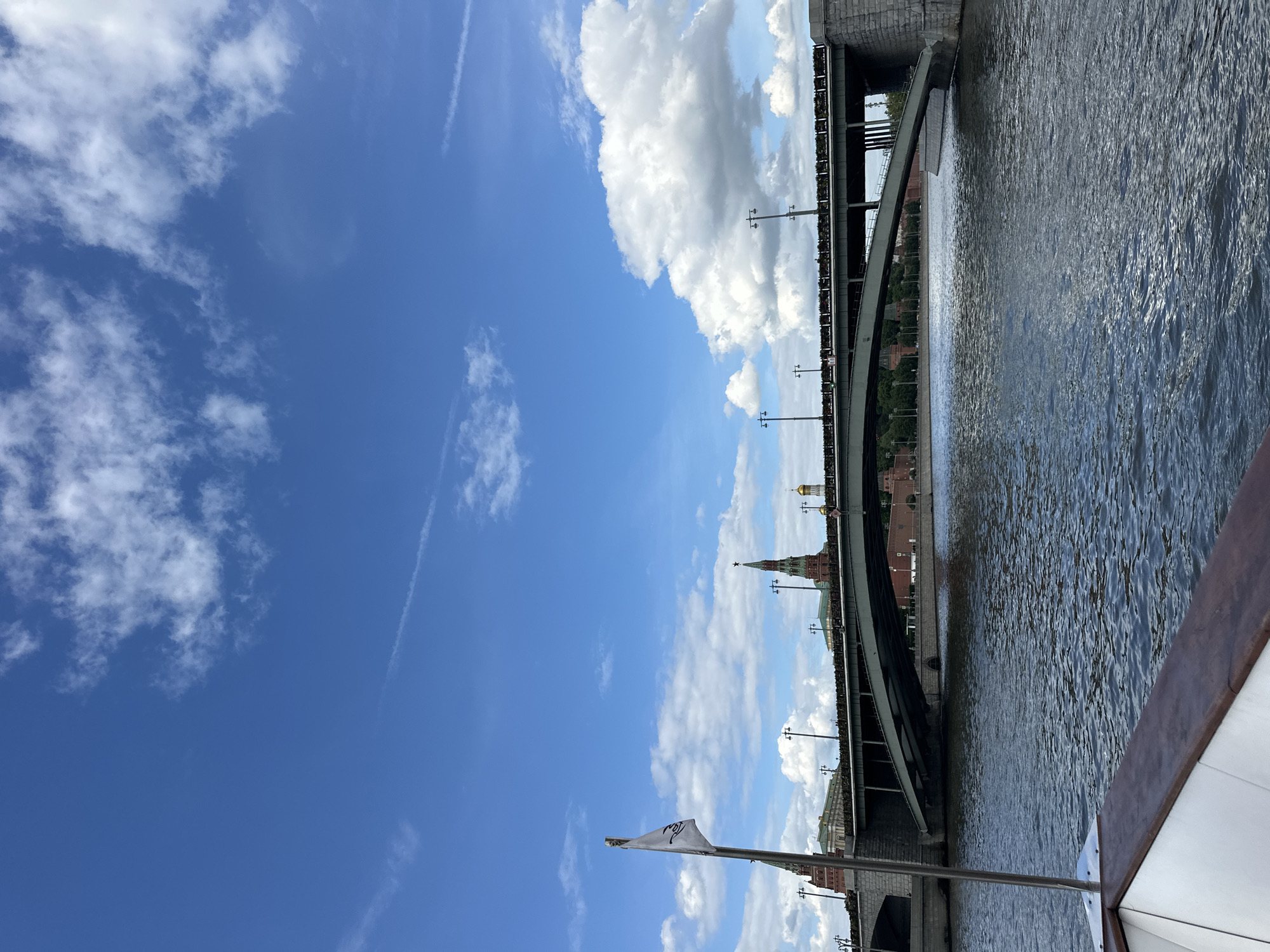 The image depicts a scene in Moscow, Russia, likely captured from a boat on the Moskva River. The view includes the Kremlin towers and walls alongside a bridge. The foreground is filled with the blue sky with some scattered clouds and the water of the river. The bridge appears to be a large overpass. The architecture in the background is consistent with Moscow landmarks. There are street lights attached to the side of the bridge.
