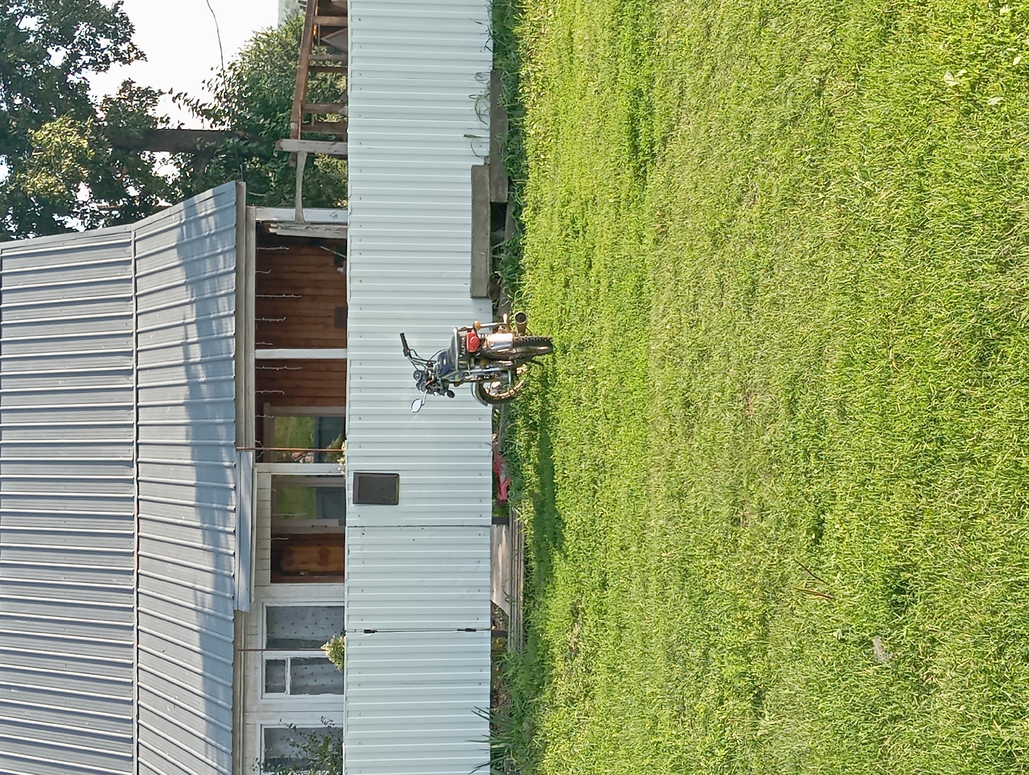In Новокопылово, Russia, a motorcycle is parked on a grassy lawn in front of a white corrugated metal fence. Behind the fence, a house with a silver metal roof and wooden-paneled porch is visible. The house has white framed windows and doors. A large tree with green leaves stands to the right of the house, and some branches of another tree are visible in the upper right corner. The sky is bright and clear. The motorcycle is dark colored with chrome accents, and the word "ALPHA" is visible on its rear. A rectangular concrete object is placed in the grass near the fence.