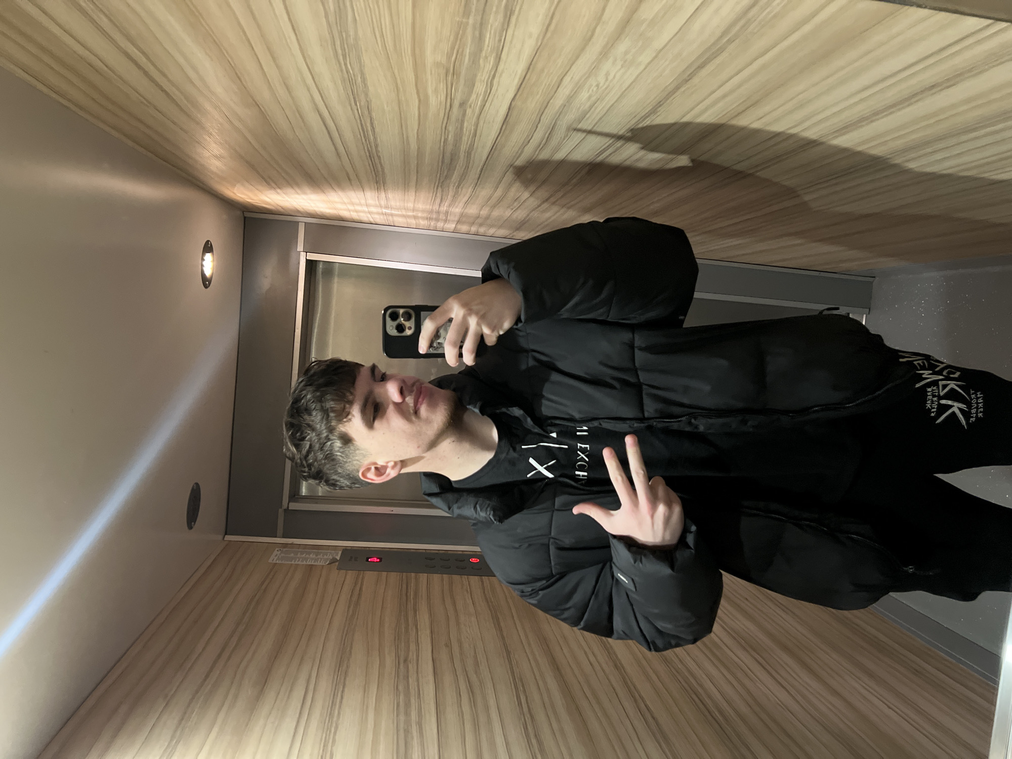 A young man is taking a selfie in an elevator. He is wearing a black puffer jacket over a black t-shirt and black pants. He is holding his phone in his right hand and making a peace sign with his left hand. The elevator has wood-paneled walls and a metal door. There is a light fixture visible on the wall behind him. The location of the elevator in the city of Sofia, Bulgaria is unknown.