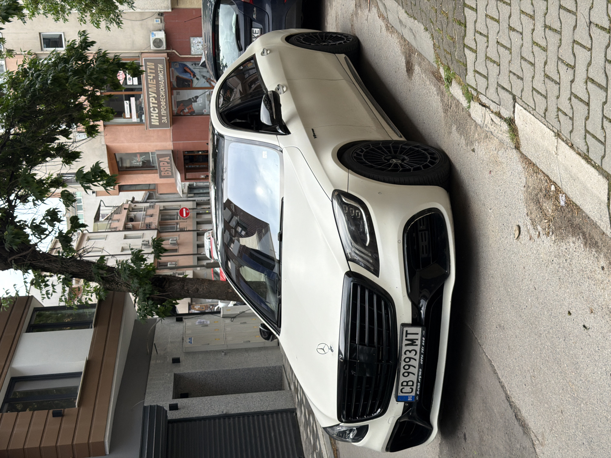 A white Mercedes-Benz sedan is prominently featured, parked at an angle on a city street in Plovdiv, Bulgaria, during daytime. The car, possibly an AMG variant with a visible badge on the front fender, has a black grille, black multi-spoke alloy wheels, and dark trim. Its Bulgarian license plate, "CB 9993 MT," is clearly visible on the front bumper. The license plate frame also displays "БОГДАН АВТО" and a phone number (0896 781 668).
To the left, a large tree with green leaves stands next to the sidewalk. In the background, several multi-story buildings line the street. One building houses a ground-floor shop with a sign in Cyrillic that reads "ИНСТРУМЕНТИ ЗА ПРОФЕСИОНАЛИСТИ" (Tools for Professionals), and another partially visible sign below it states "ВЯРА" (Faith). Air conditioning units are attached to the building facades. A red circular "No Entry" traffic sign is also visible further down the street. The sidewalk is paved with interlocking concrete blocks, and the street is asphalt. No people are visible, and the scene depicts a typical urban street with a parked vehicle under clear or partly cloudy skies.