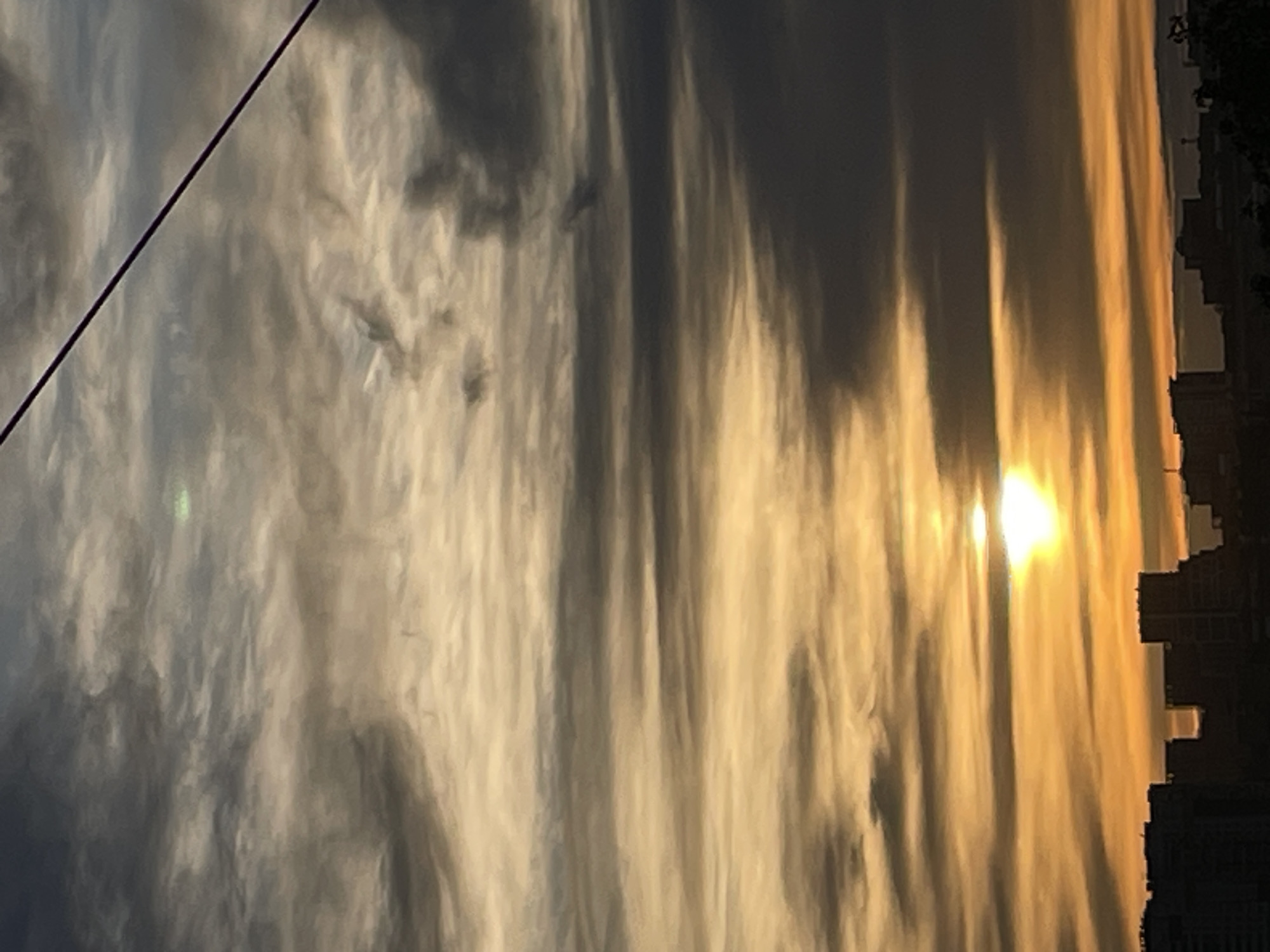 The image depicts a bright light source, likely the sun, shining through a sky with prominent cloud formations. Vertical streaks of light and shadow are visible across the frame. In the lower right corner, the silhouette of buildings, presumably part of a city skyline, can be discerned. A wire stretches diagonally across the left side of the image. The image is likely taken in Izhevsk, Russia, given the prompt, though visual cues confirming this are absent.