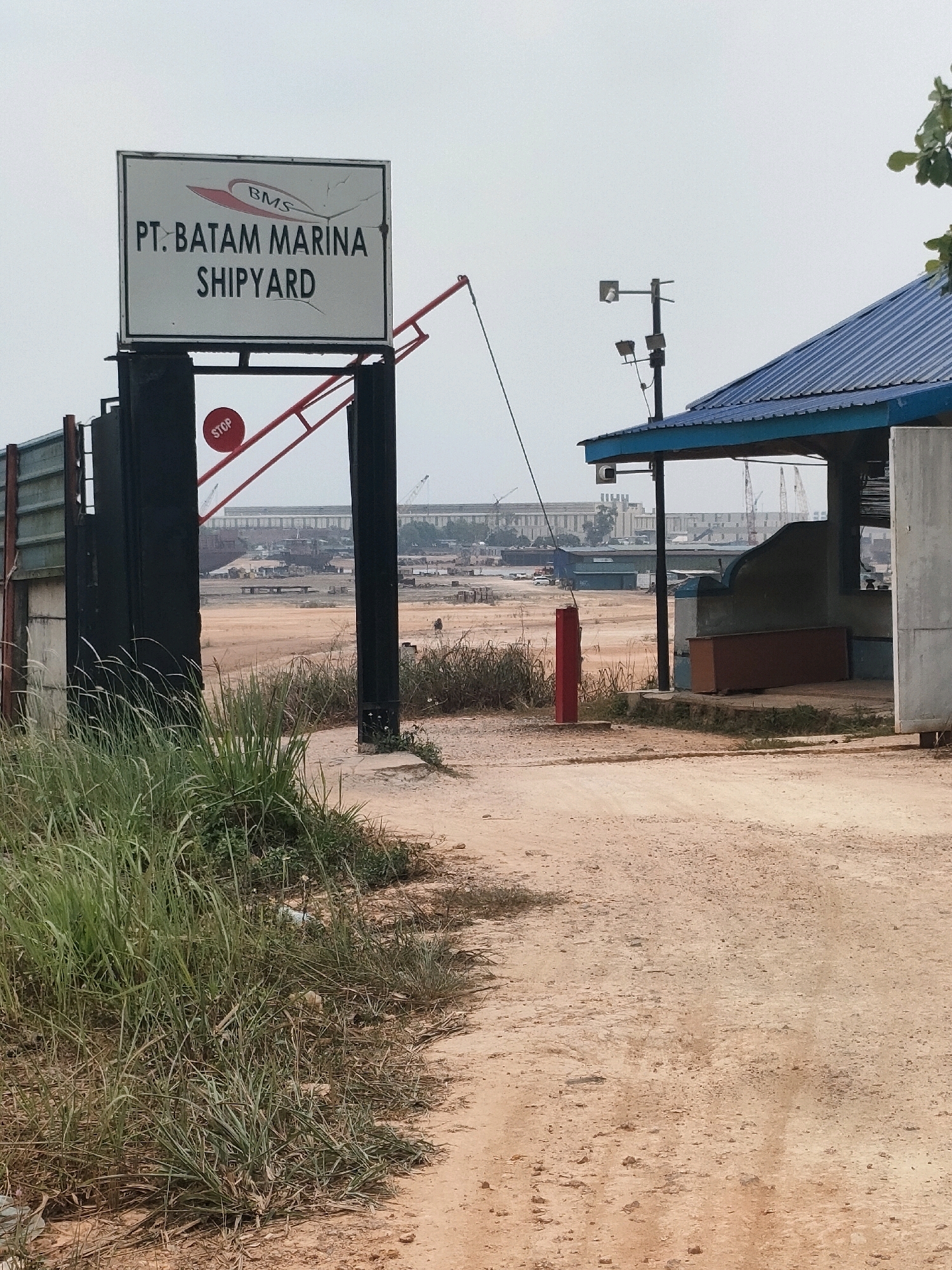 A sign displaying "BMS PT. BATAM MARINA SHIPYARD" hangs on a black metal post at the entrance to a shipyard. A red stop sign is mounted on a red boom gate. A dirt road leads into the facility, which features a blue-roofed building with lights and a security camera. In the background, industrial structures, cranes, and ships are visible, indicating a busy port or shipbuilding operation. The sky is overcast. The location is the PT. Batam Marina Shipyard in Batam, Indonesia.