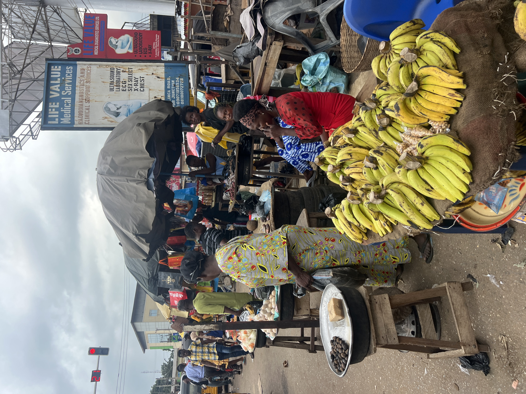 This image captures a bustling outdoor market scene in Nkpor, Nigeria. Several people are visible, with a focus on individuals engaged in commerce. A woman in a vibrantly patterned dress stands behind a stall displaying bananas, eggs, and what appear to be nuts. Another woman in a red dress with black polka dots is seated nearby. The market is set up along a street, with various stalls and temporary structures contributing to a lively, somewhat cluttered environment.
Prominent signage for "LIFE VALUE Medical Services" is visible, advertising services such as ultrasound, laboratory tests, ECG, and X-ray. This suggests the market is located near a medical facility or that medical services are being advertised to the local community. The presence of a traffic light with a red signal indicates that this is an active intersection. The weather appears to be overcast, with a cloudy sky.
The overall impression is one of a typical day in a busy Nigerian market, with vendors and customers interacting amidst the sale of produce and other goods. The people's expressions are mostly neutral, suggesting the focus is on the transactions and daily activities rather than strong emotional displays. The ground is paved, and there are various items for sale, including a large quantity of bananas. The scene is rich with the details of everyday life in a Nigerian urban setting.