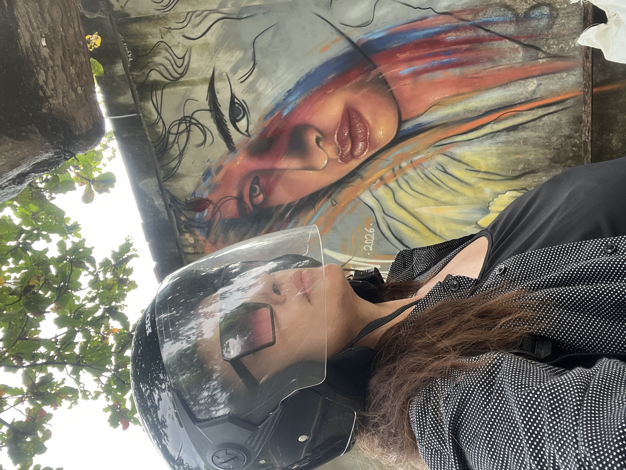This media file features a person wearing a motorcycle helmet and a polka-dot patterned jacket, partially obscuring a vibrant mural. The mural, likely painted on a concrete wall, depicts a stylized face with flowing, colorful hair rendered in shades of red, orange, blue, and yellow. The person's face is visible through the helmet's clear visor, their gaze directed downwards. The helmet itself has "HONDA" subtly embossed on it.
The scene is outdoors, with lush green foliage and tree branches visible in the upper left and right portions of the frame, suggesting a tropical or subtropical environment. The lighting indicates it is daytime, possibly overcast or partially shaded by the trees. The overall impression is one of casual exploration or a momentary pause during travel. The mural's artistic style and the presence of the motorcycle helmet suggest an urban or semi-urban setting in Kuta, Indonesia, known for its street art and bustling atmosphere. A small, barely legible inscription "2026." is visible on the mural.