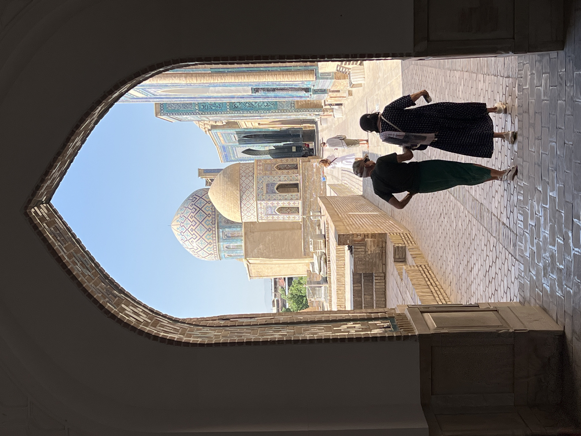 This image captures a view through a large arched entryway onto a courtyard of the Registan complex in Samarkand, Uzbekistan. The courtyard is paved with light-colored cobblestones. In the foreground, two individuals are walking away from the viewer, with one wearing a dark t-shirt and green pants, and the other in a dark patterned dress. Behind them, two more individuals, one in a white dress and the other in a light gray outfit, are walking further into the complex.
Dominating the background are several impressive structures adorned with intricate mosaic tilework. The most prominent is a turquoise and blue domed building with geometric patterns, likely a madrasah or mosque. Adjacent to it are other buildings with similar blue and white tile decorations, featuring arched windows and tall minarets. The sky above is clear and blue, suggesting a sunny day. The architecture indicates a style typical of Timurid-era Central Asian Islamic complexes.