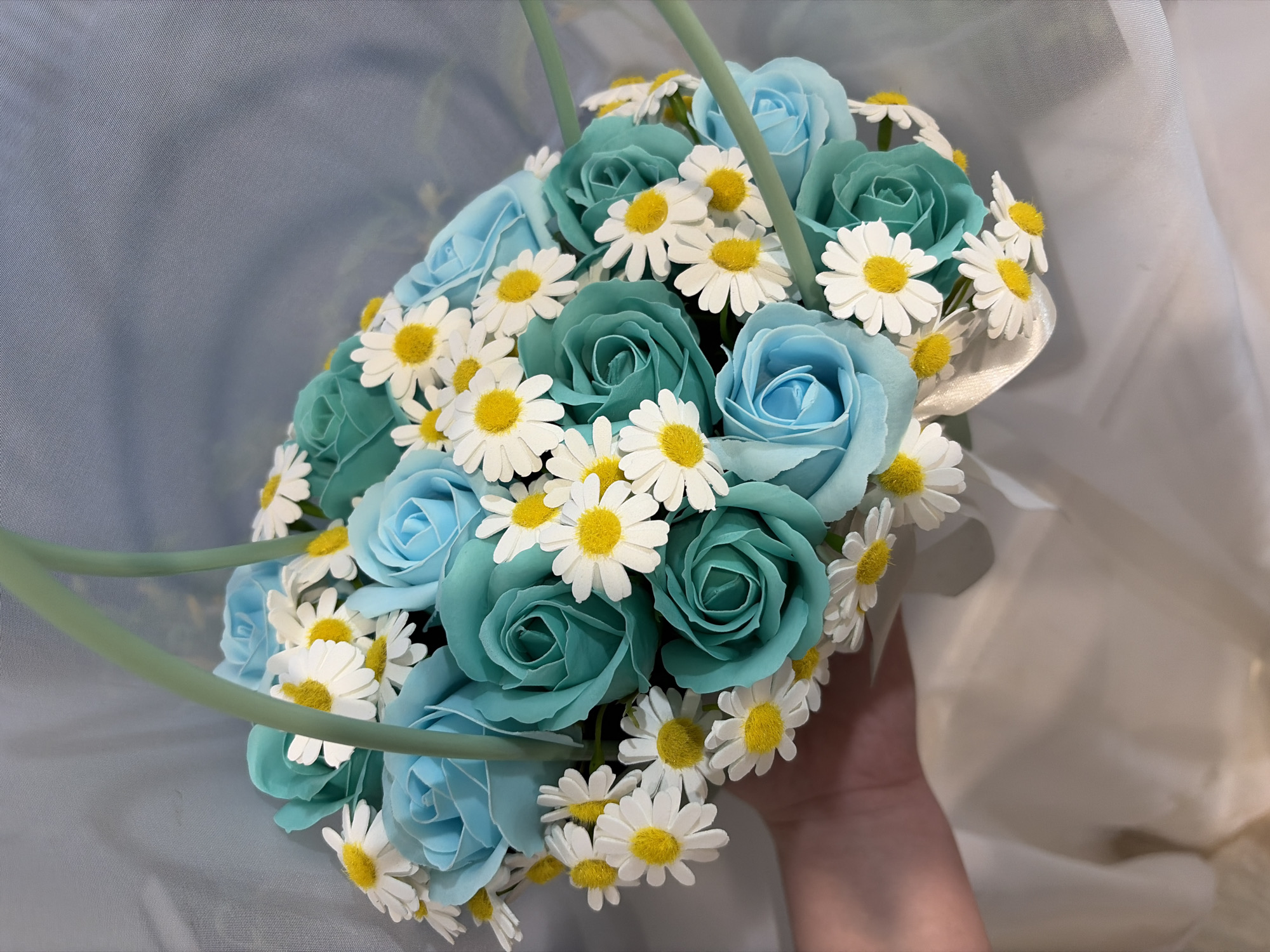 The image features a close-up of a bouquet of artificial flowers, comprised of roses in shades of turquoise and light blue, interspersed with small daisies. The bouquet is held in a hand against a backdrop of draped white fabric. The arrangement includes a light green handle. The location details are not discernible from the image.