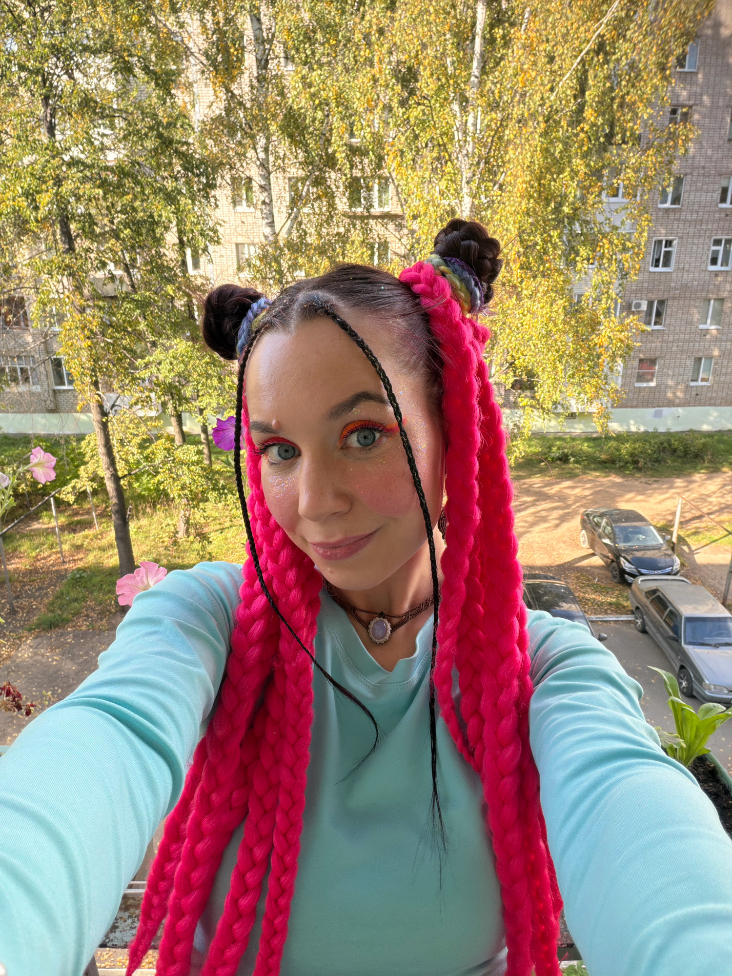 A close-up selfie of a woman with bright pink braided hair and glitter makeup is taken outdoors in Chaikovsky, Russia. The woman is wearing a light blue long-sleeved shirt and a necklace with a stone pendant. She has two buns in her hair, adorned with rainbow-colored accents. In the background, there is a brick apartment building, trees with yellowing leaves indicating autumn, and parked cars. Pink and purple petunias are visible on a balcony in the foreground. The lighting suggests it is daytime.