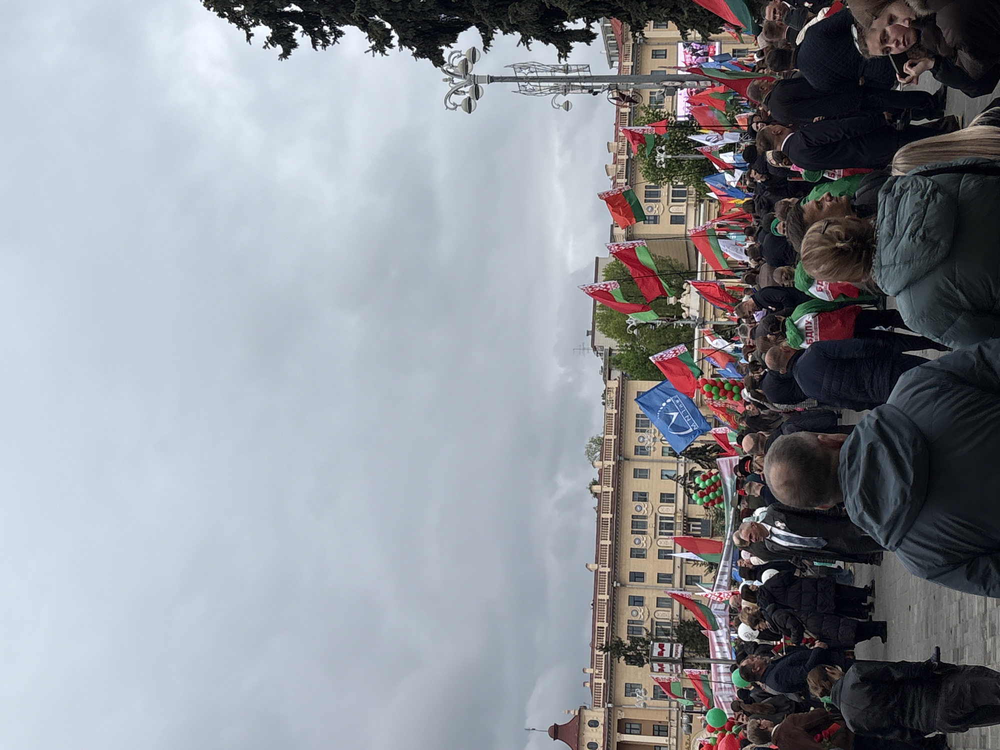 The image shows a crowd of people gathered in a public square in Minsk, Belarus. The people are holding various flags, primarily the red and green flag of Belarus. There are also some blue and white flags visible. The crowd is dense, and people appear to be standing close together. In the background is a large building with many windows. The sky is overcast and gray. A decorative lamppost is visible among the crowd and flags. Trees are visible at the top of the frame. The composition suggests a civic event or demonstration. The environment is an urban setting with buildings and infrastructure.