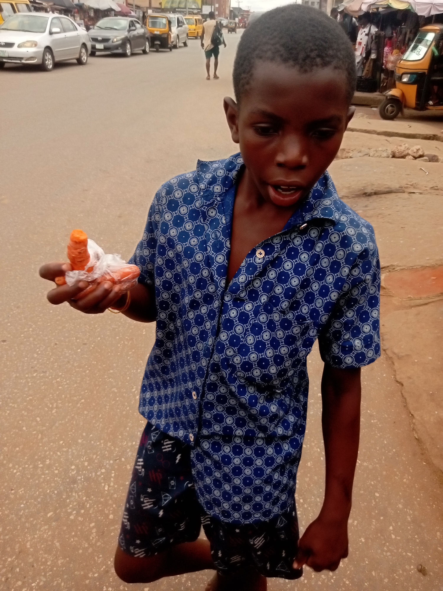 A young, dark-skinned boy stands on a street in Nkpor, Nigeria. He is wearing a blue, patterned short-sleeved shirt and dark shorts with white designs. He is holding a small plastic bag containing peeled carrots in his left hand. His mouth is open slightly, as if speaking or surprised. The street is paved and appears somewhat worn. In the background, there are various vehicles, including cars and a three-wheeled vehicle (keke), as well as other people. There are buildings and stalls along the side of the road. The overall impression is of a bustling street scene.
