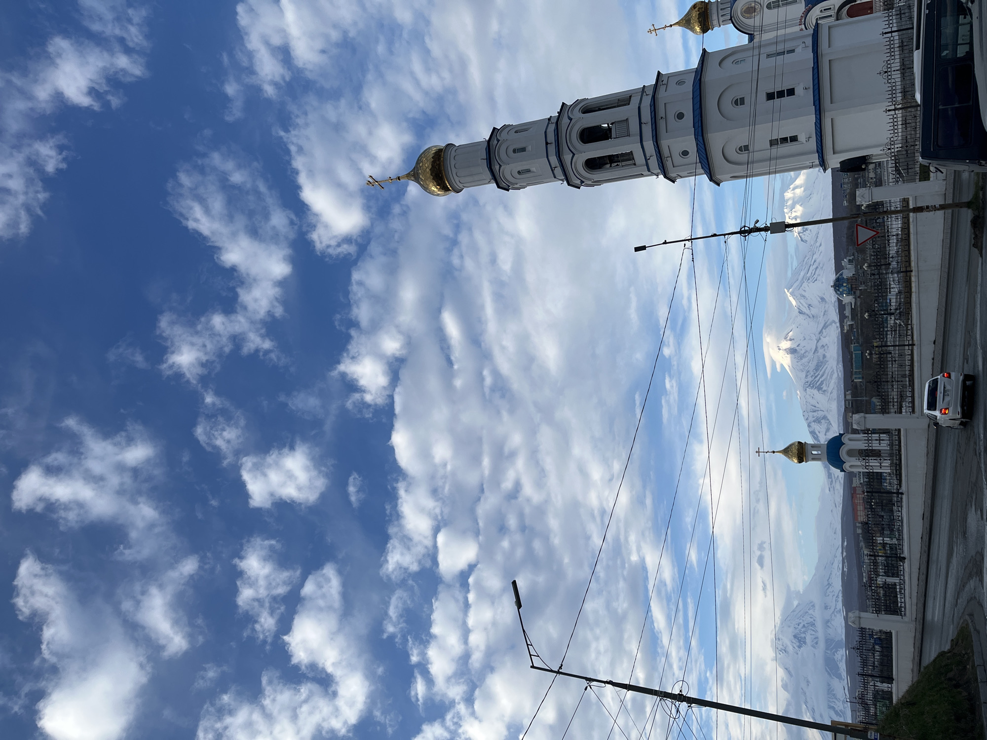 The image shows a street scene in Petropavlovsk-Kamchatsky, Russia, featuring two Eastern Orthodox churches. The lower church is the Holy Trinity Cathedral. The photograph is taken from a low angle looking upwards. The bell tower of the Holy Trinity Cathedral extends upwards and is oriented vertically. The sky behind the buildings is blue and filled with clouds. In the background, snow-covered mountains are visible. A street with vehicles, including a white car, is present on the right side of the image. Power lines and street lights are visible in the foreground.