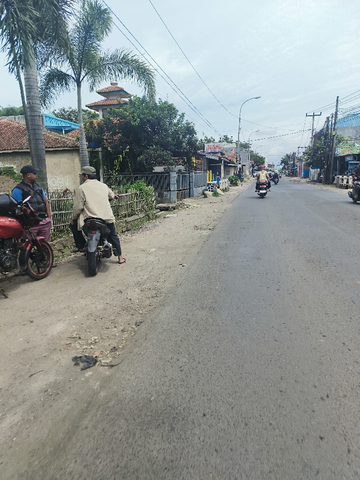 The image depicts a street scene in Ciparay, Indonesia (latitude: -7.024, longitude: 107.689). Two men are standing on the shoulder of the paved road, each with a motorcycle. Both motorcycles are parked. The man on the left is wearing a dark shirt, dark pants, and a helmet. He is holding a black bag on his left shoulder. The man on the right is wearing a beige shirt and beige pants. There are numerous buildings along the street, with small shops, homes, and some vegetation. The image is taken from the middle of the road, facing the direction of oncoming traffic. There are several power lines overhead, and a street light is visible in the distance. The asphalt road is dry and relatively empty, with only a single motorcycle traveling in the distance. The sky is overcast and gray.