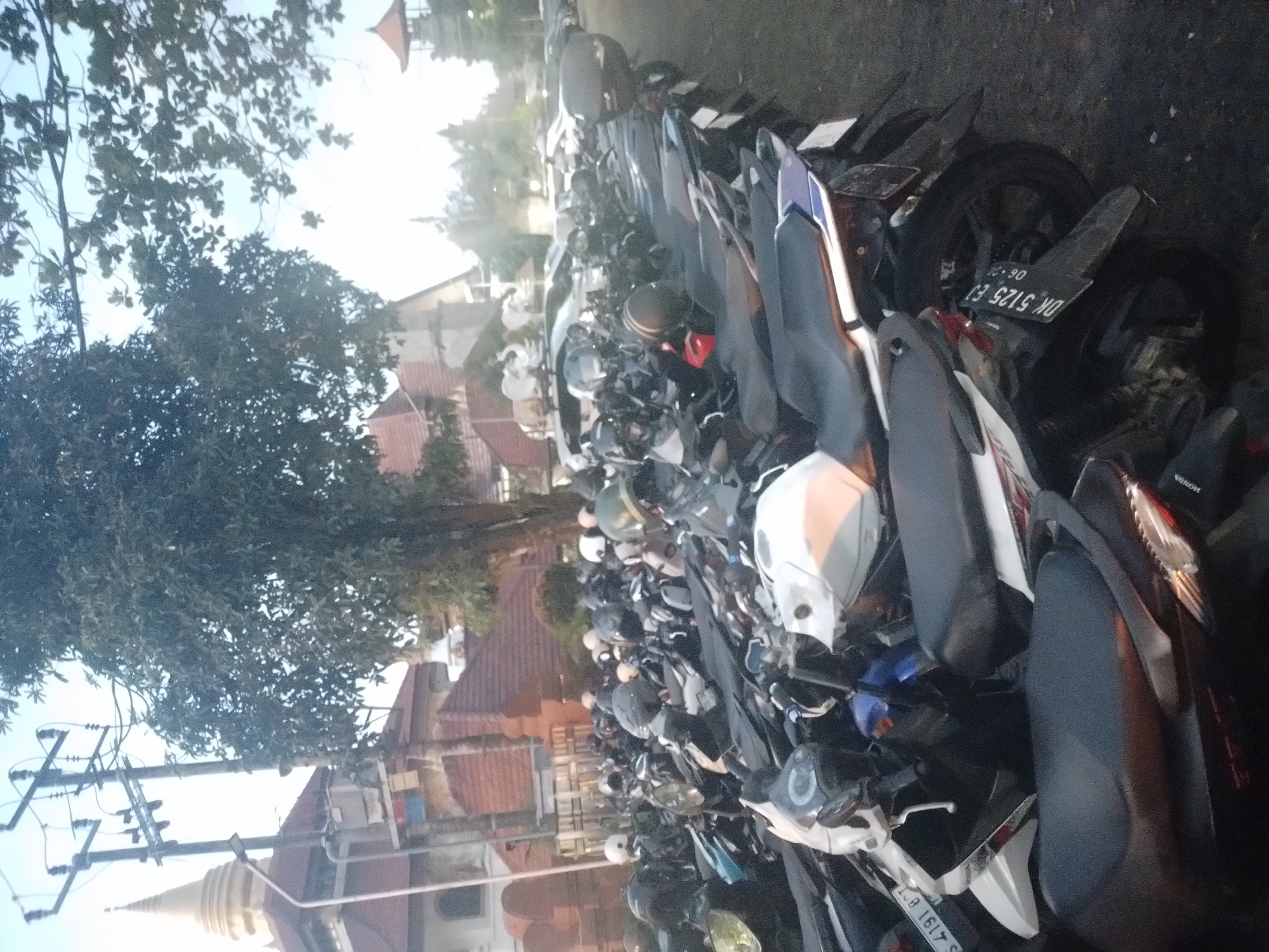 The image depicts a dense motorcycle parking area in Kuta Selatan, Indonesia. A large number of motorcycles are parked closely together. Buildings with reddish-brown roofs are visible in the background. A tree and power lines are also present.