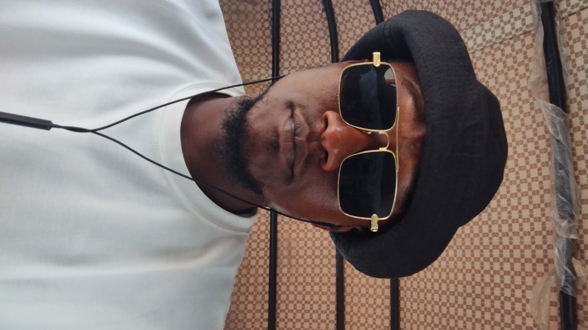 A man with dark skin and a beard is lying on his back on a surface with a checkered pattern. He is wearing a white shirt, a black hat, and gold-rimmed sunglasses. He also has earbuds around his neck. A dark, slender object wrapped in plastic is partially visible on the right side of the image. The image was taken at Jos, Nigeria.